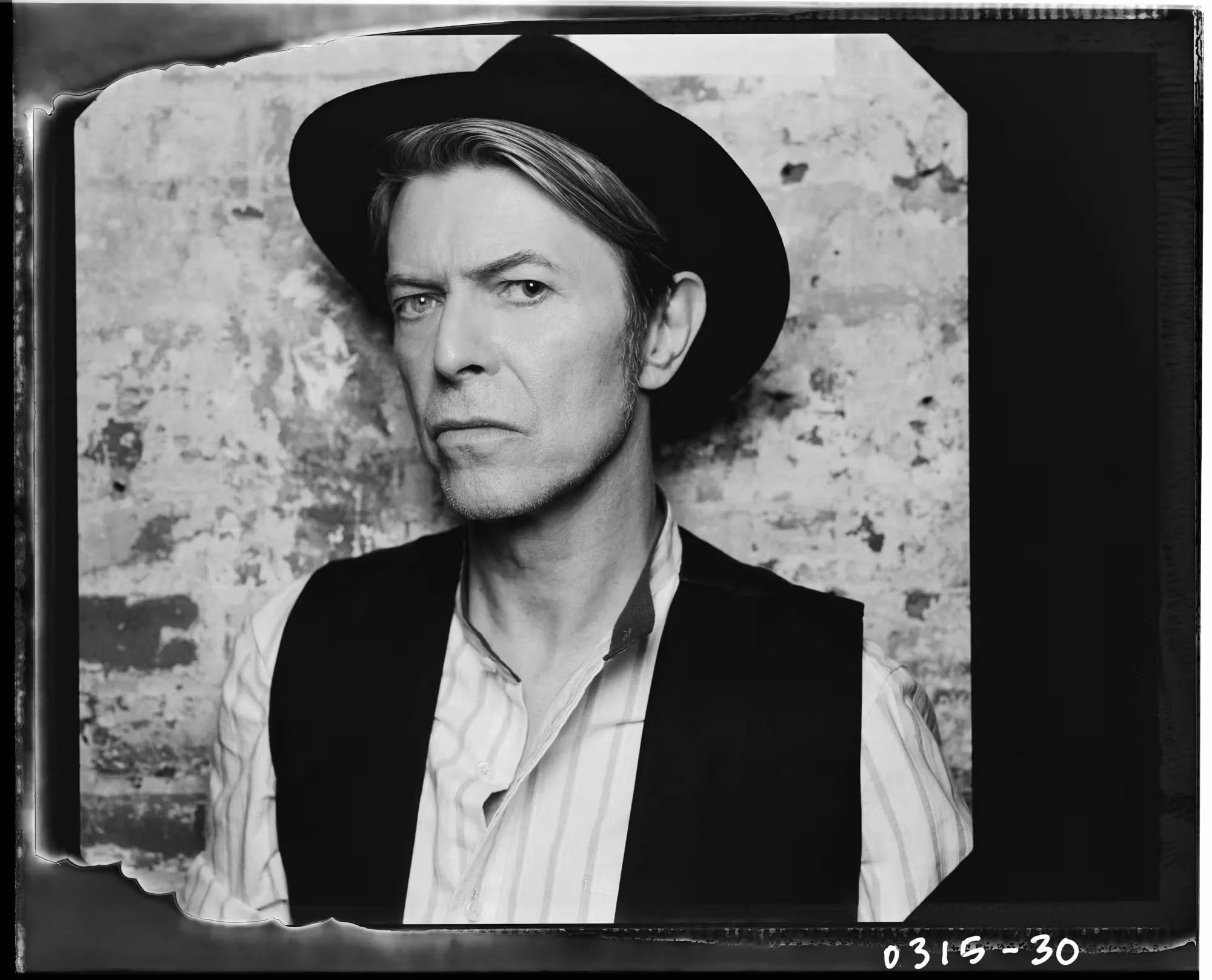 Black and white portrait of a person wearing a brimmed hat and vest, standing against a textured wall. They have a serious, contemplative expression.