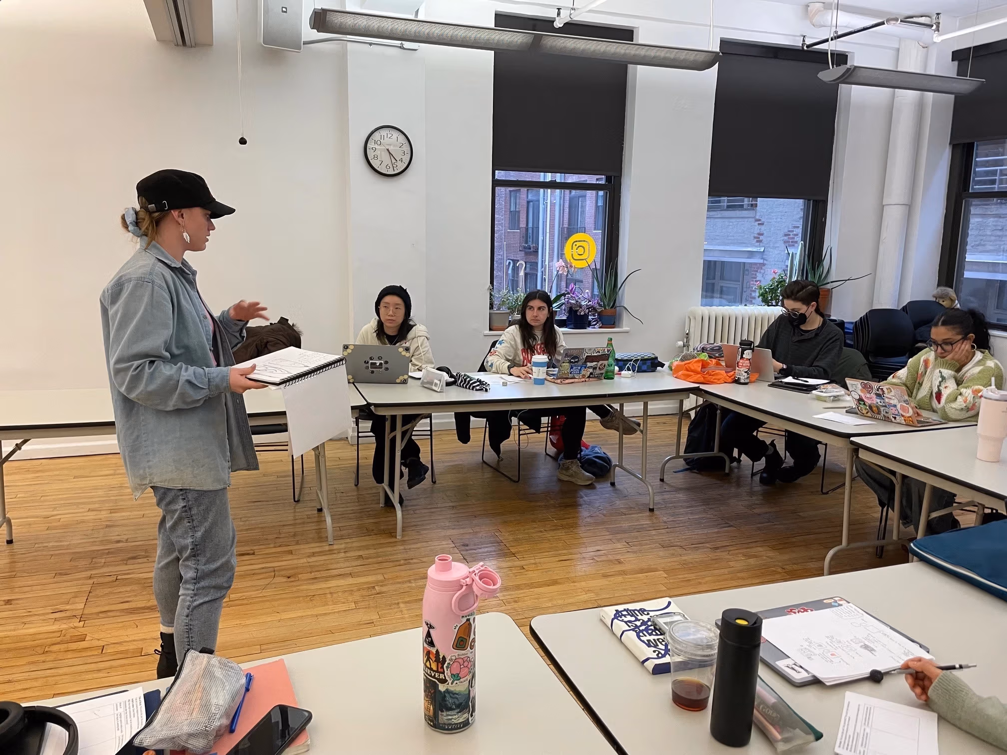 A student in denim stands in front of her classmates sitting in a u-shape arrangement. The student standing is addressing her classmates with a sketchbook in hand.