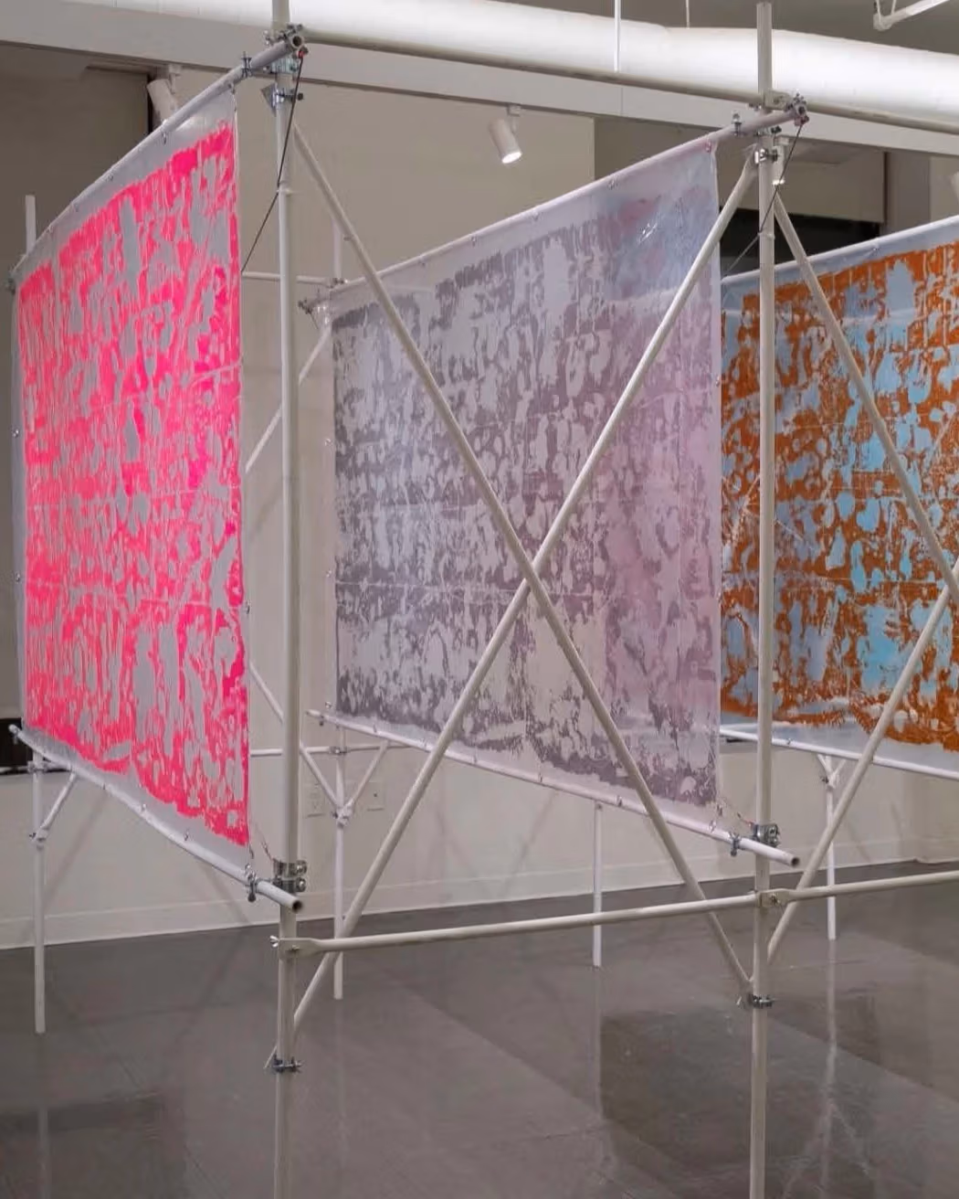 5 large rectangular painted fabric sheets stretched and spaced equally within a steel piped structure. This structure holds all five in a row. Each fabric sheet shows amorphic shapes. Each panel displays two-three different colors. Each sheet is a different color scheme.