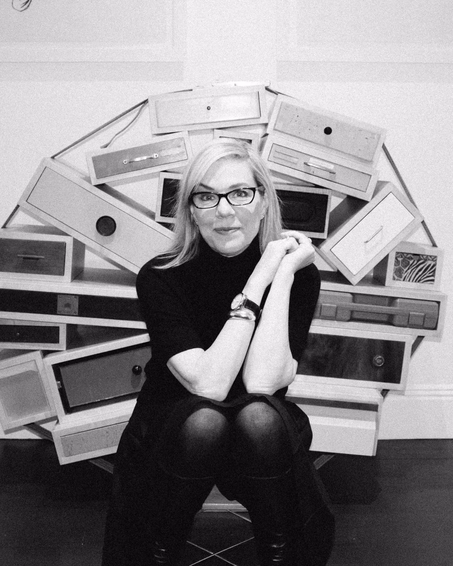 A woman with glasses and a black turtleneck sits in front of this sculpture that looks like a bunch of drawers bound together in a circle.