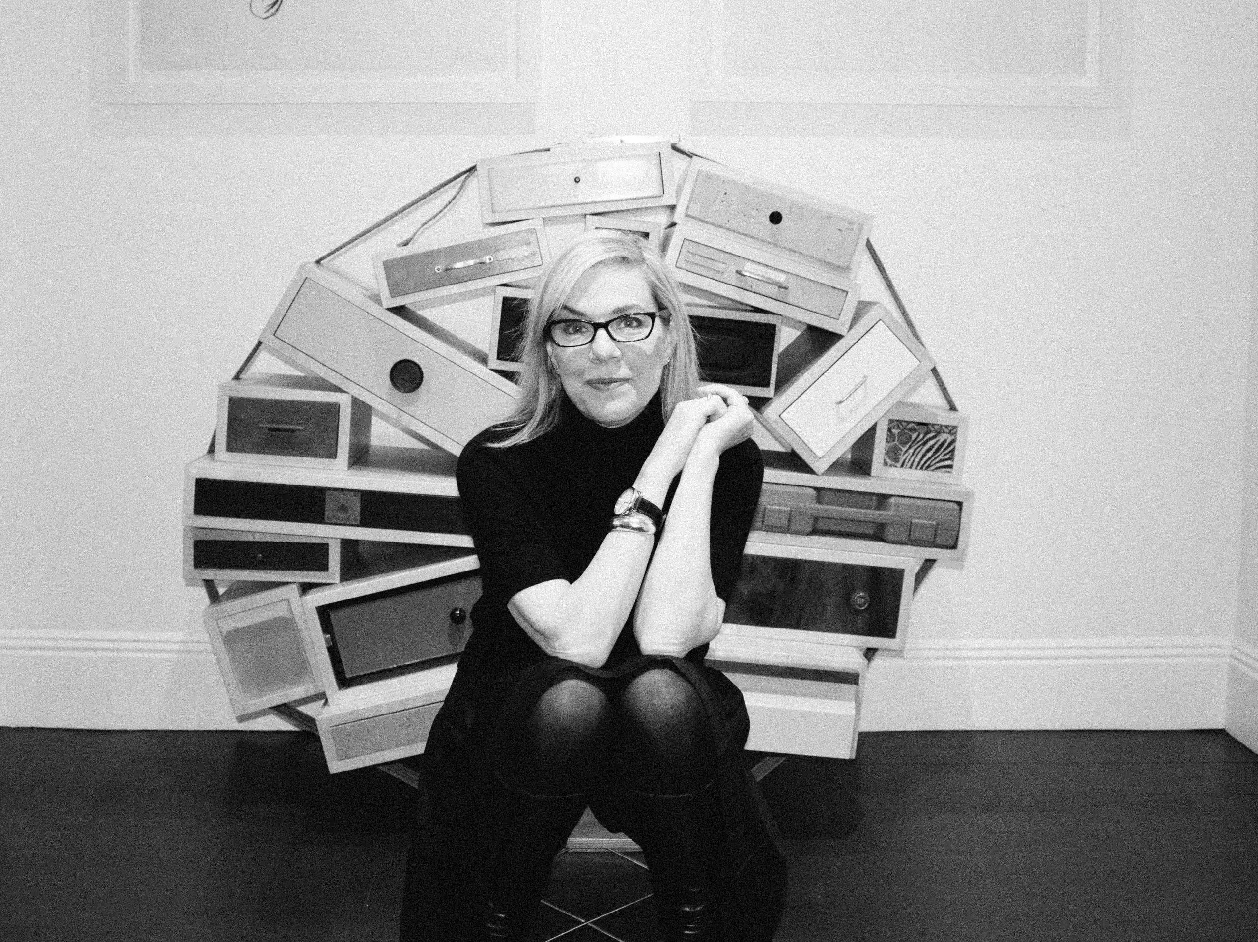 A woman with glasses and a black turtleneck sits in front of this sculpture that looks like a bunch of drawers bound together in a circle.