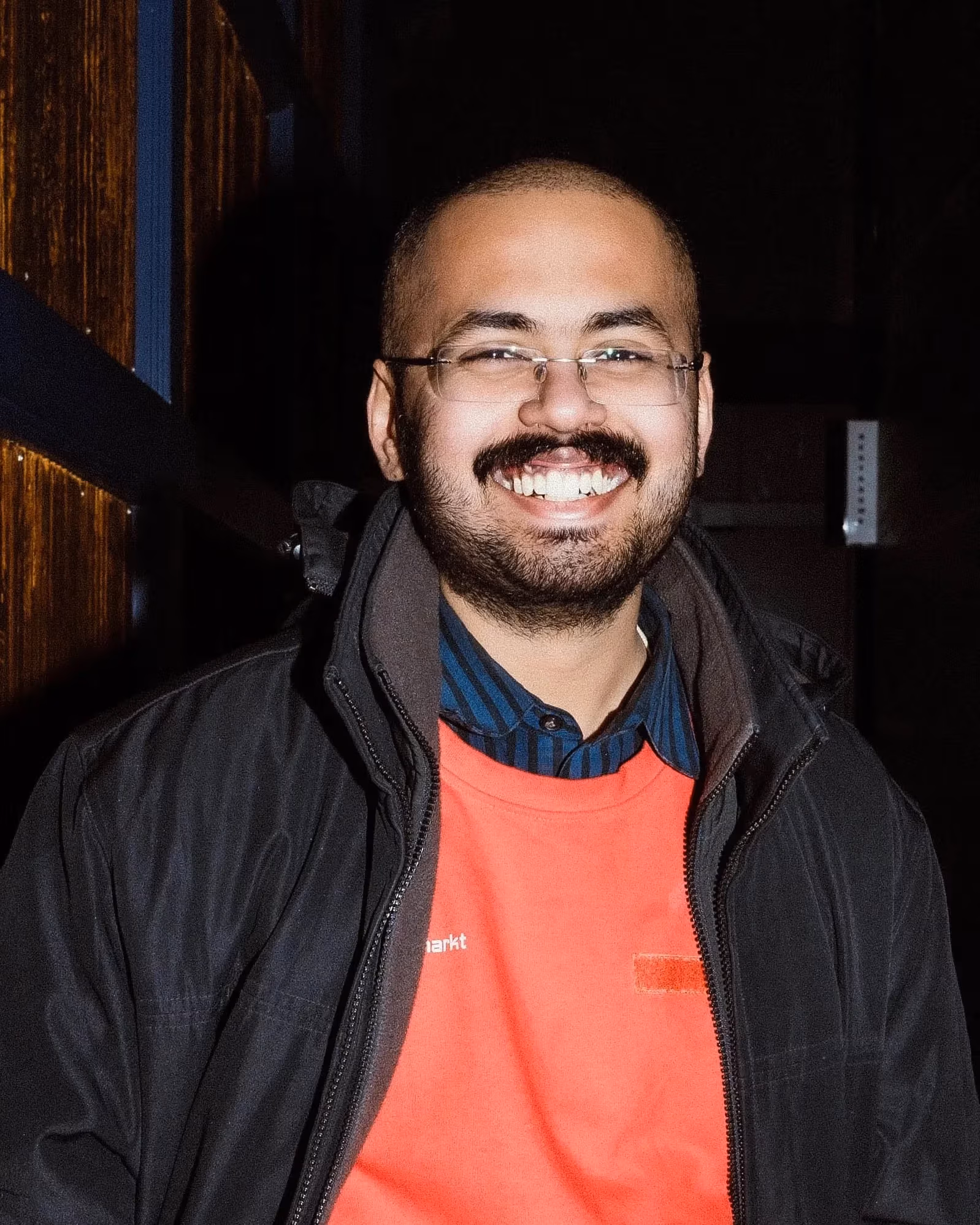 A man with a buzz cut with a dark mustache smiles at the camera wearing a red shirt and dark jacket. 