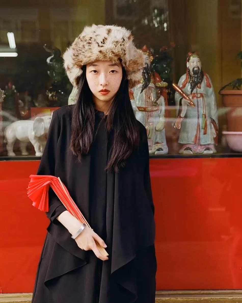 Portrait of a young woman with a big fuzzy hat posing in front of a glass window displaying porcelain sculptures. 