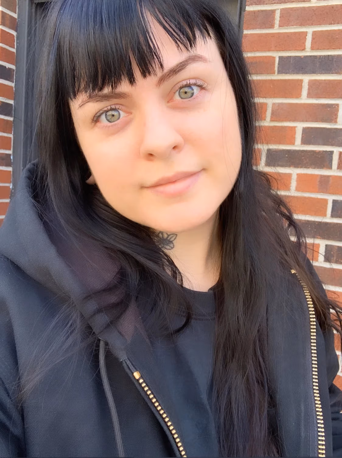 A headshot of Erin M. Riley. Erin is wearing a black shirt and black jacket. She is looking into the camera and smiling. In the background is a brick wall.