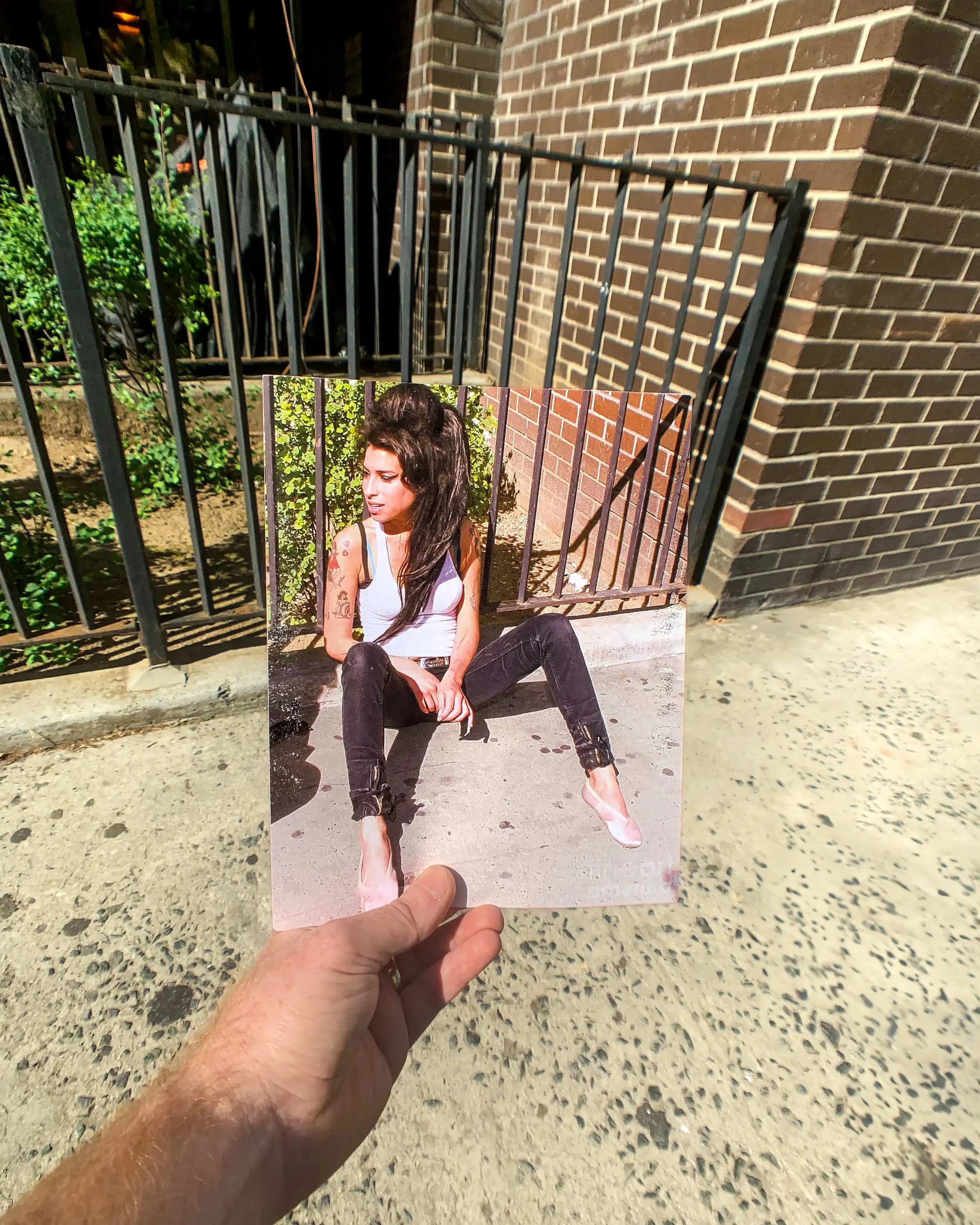 A hand holds a photo of a person sitting on a sidewalk against a brick wall and metal fence. The background aligns with the photo, blending real and image.