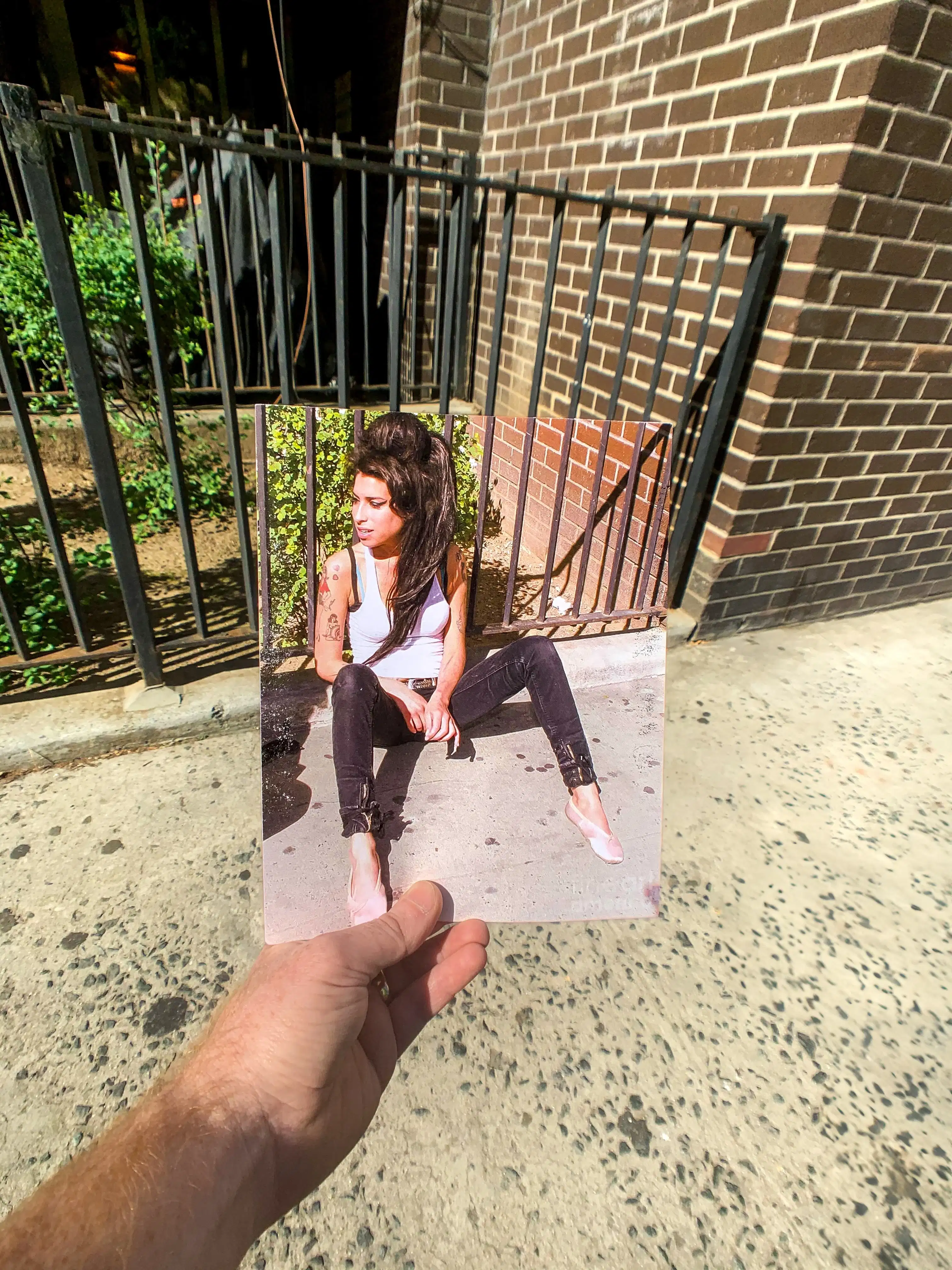 A hand holds a photo of a person sitting on a sidewalk against a brick wall and metal fence. The background aligns with the photo, blending real and image.