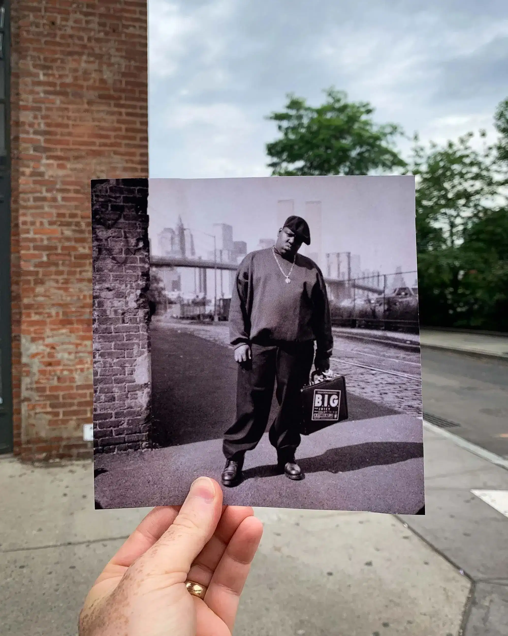 A person holds a black-and-white photo against an urban backdrop. The photo features a person in a cap and sweater, exuding a relaxed, contemplative mood.