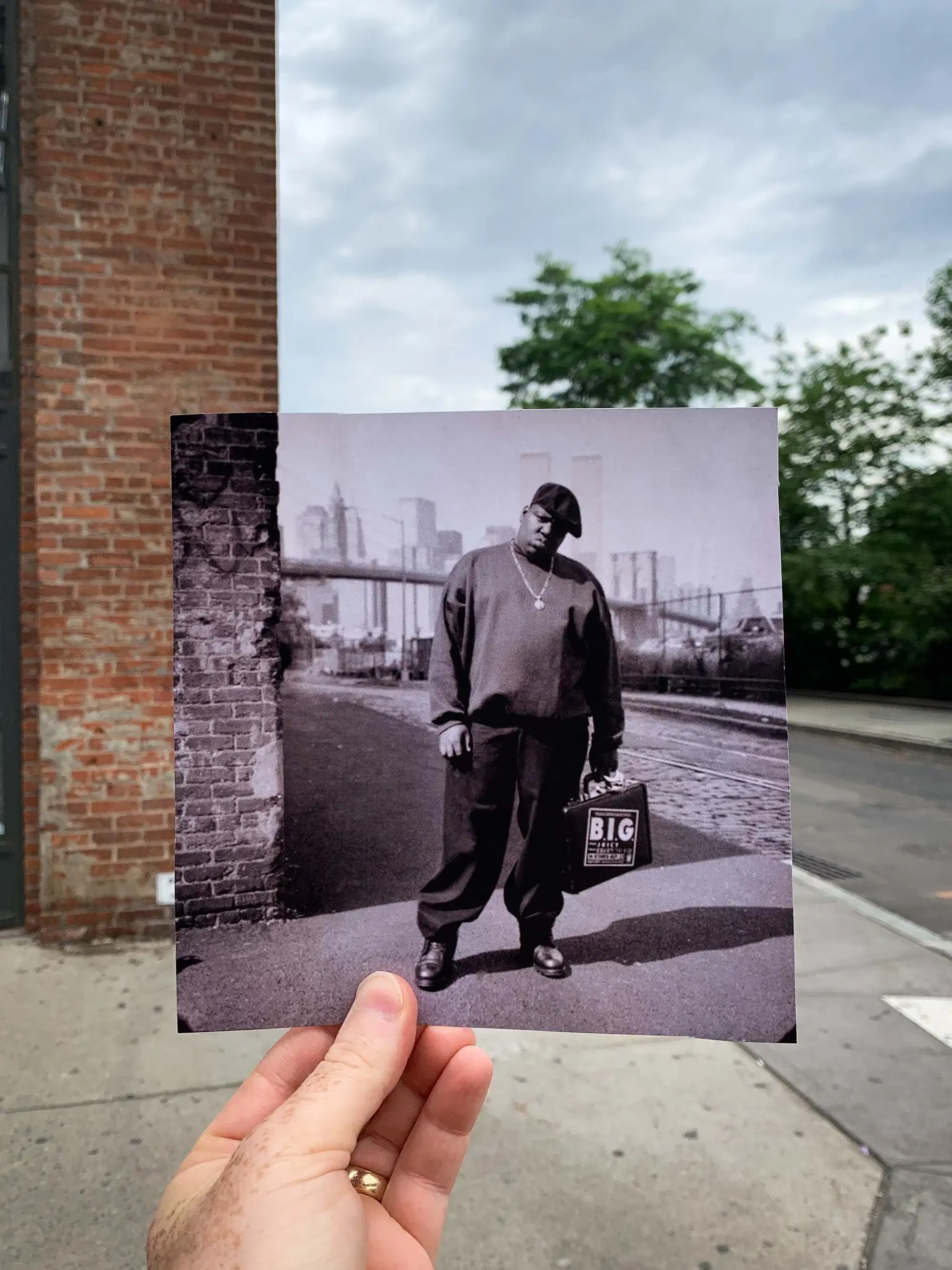 A person holds a black-and-white photo against an urban backdrop. The photo features a person in a cap and sweater, exuding a relaxed, contemplative mood.