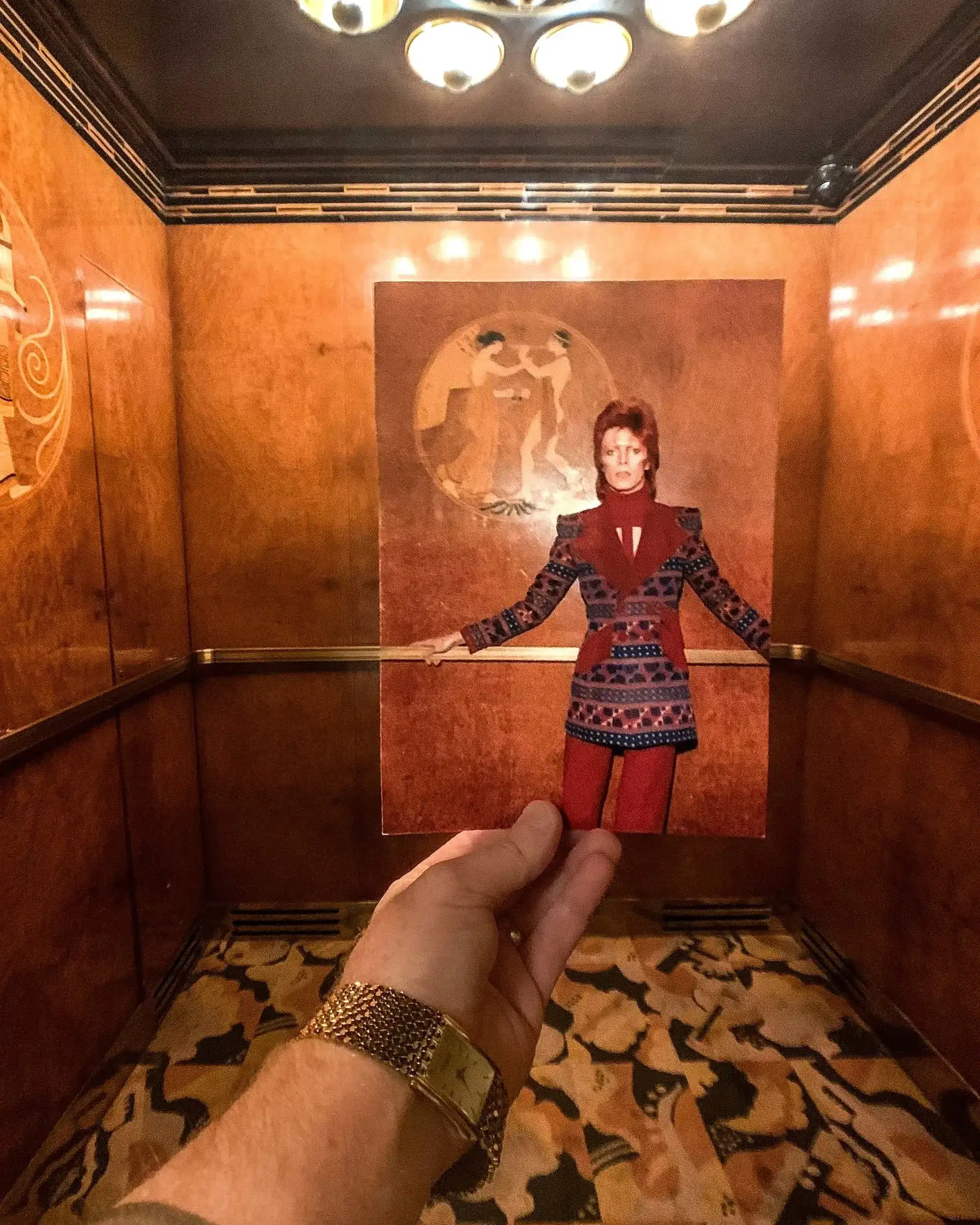 A hand holds a vintage photo in an ornate elevator. The photo features a person in a colorful, patterned outfit matching the elevator's stylish decor.