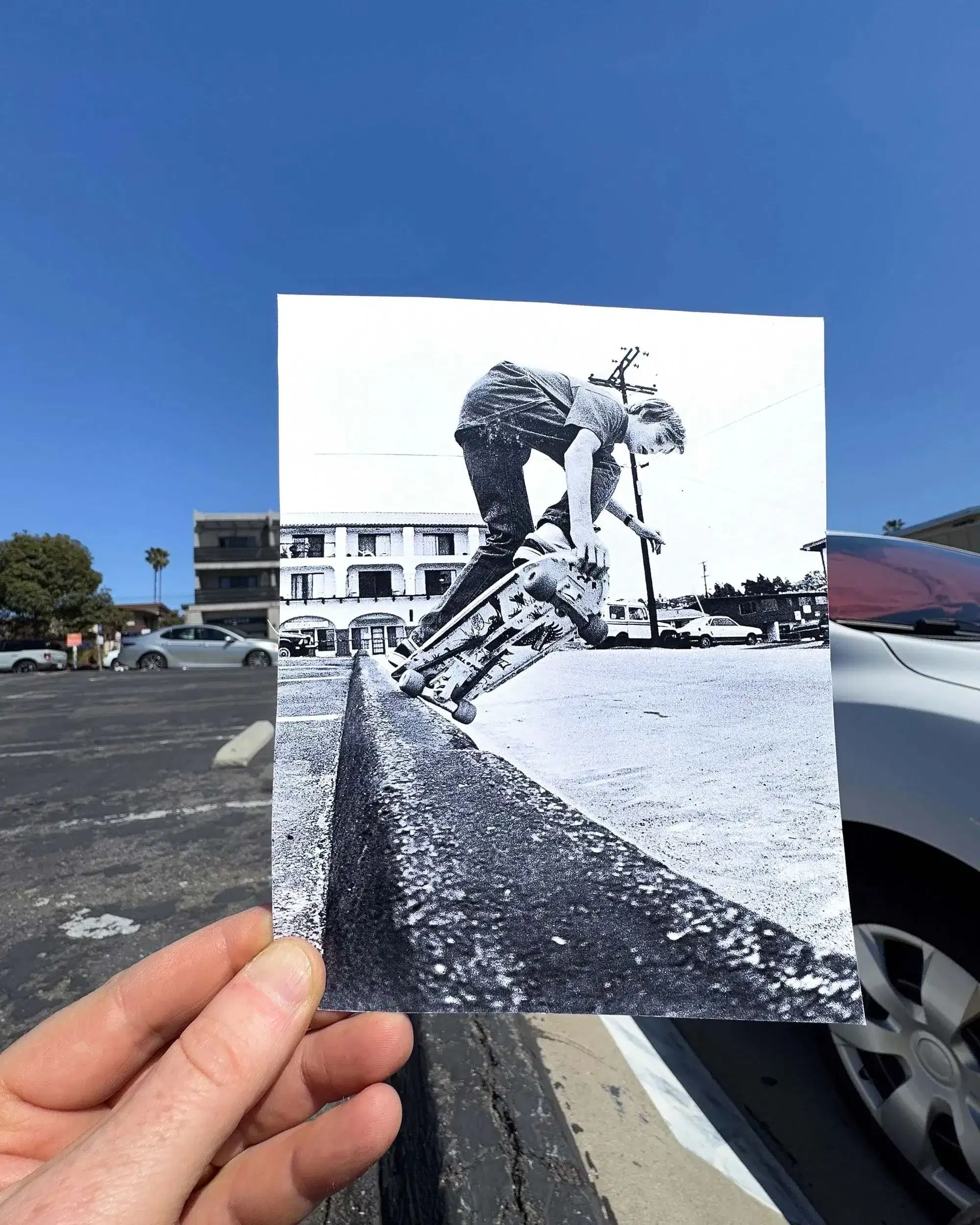 A hand holds a black-and-white photo of a skateboarder mid-trick on a curb. The background shows cars and a building under a clear, blue sky.