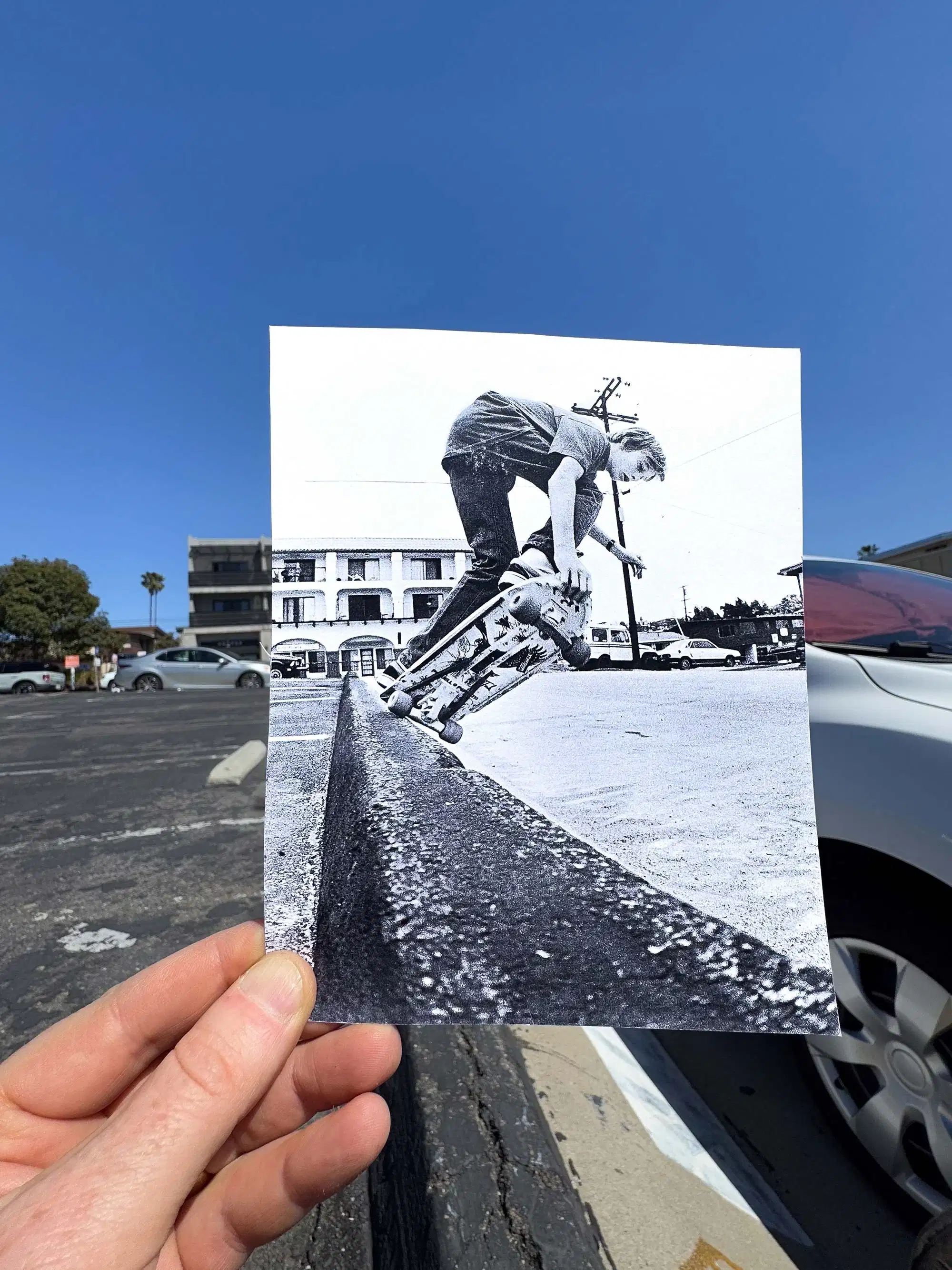 A hand holds a black-and-white photo of a skateboarder mid-trick on a curb. The background shows cars and a building under a clear, blue sky.