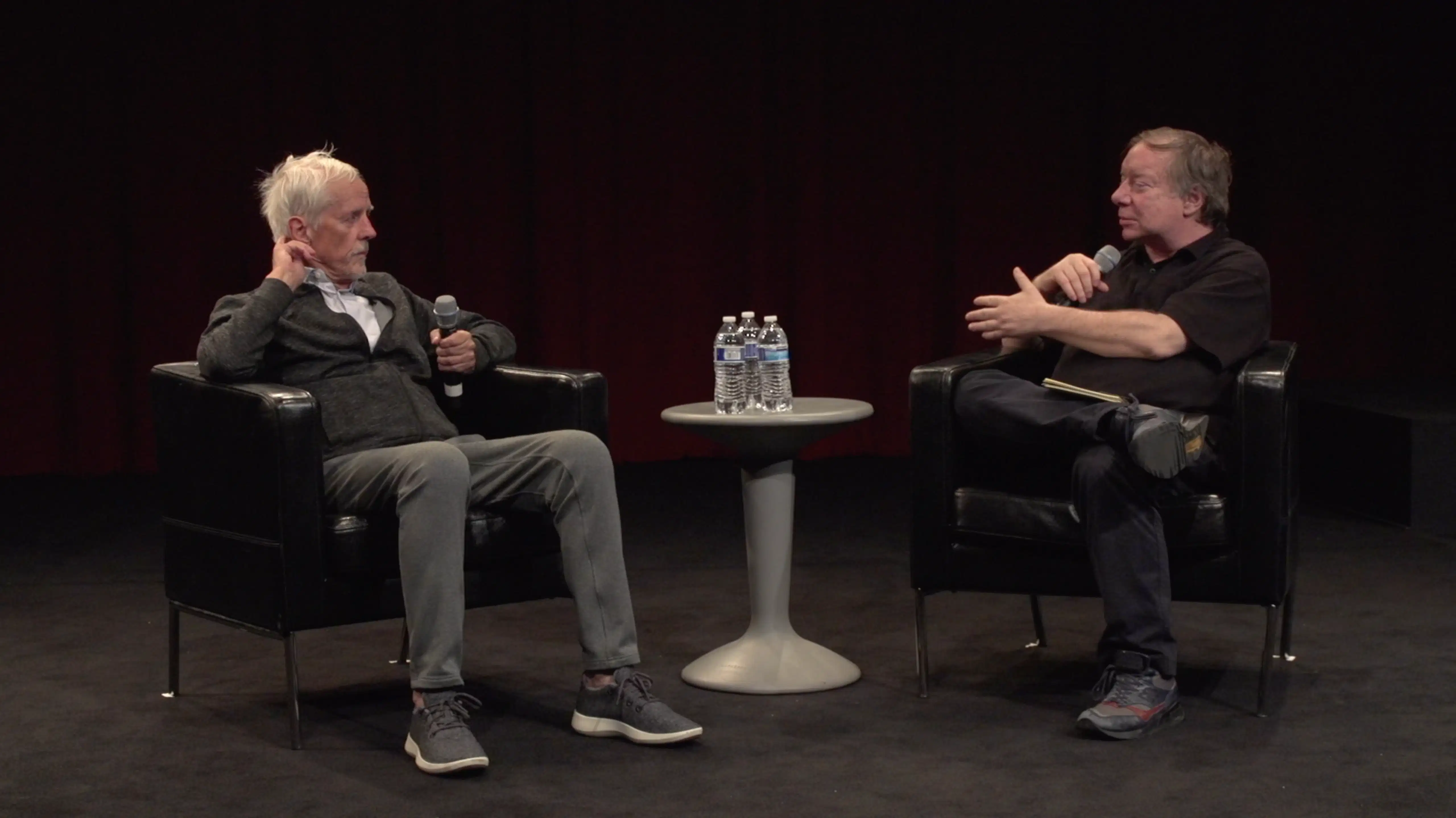 A color photograph of two people sitting in chairs on a carpeted floor, a table holding three unopened bottles of water between them. Each person is holding a microphone and the person on the right is gesturing and talking while the person on the left listens.