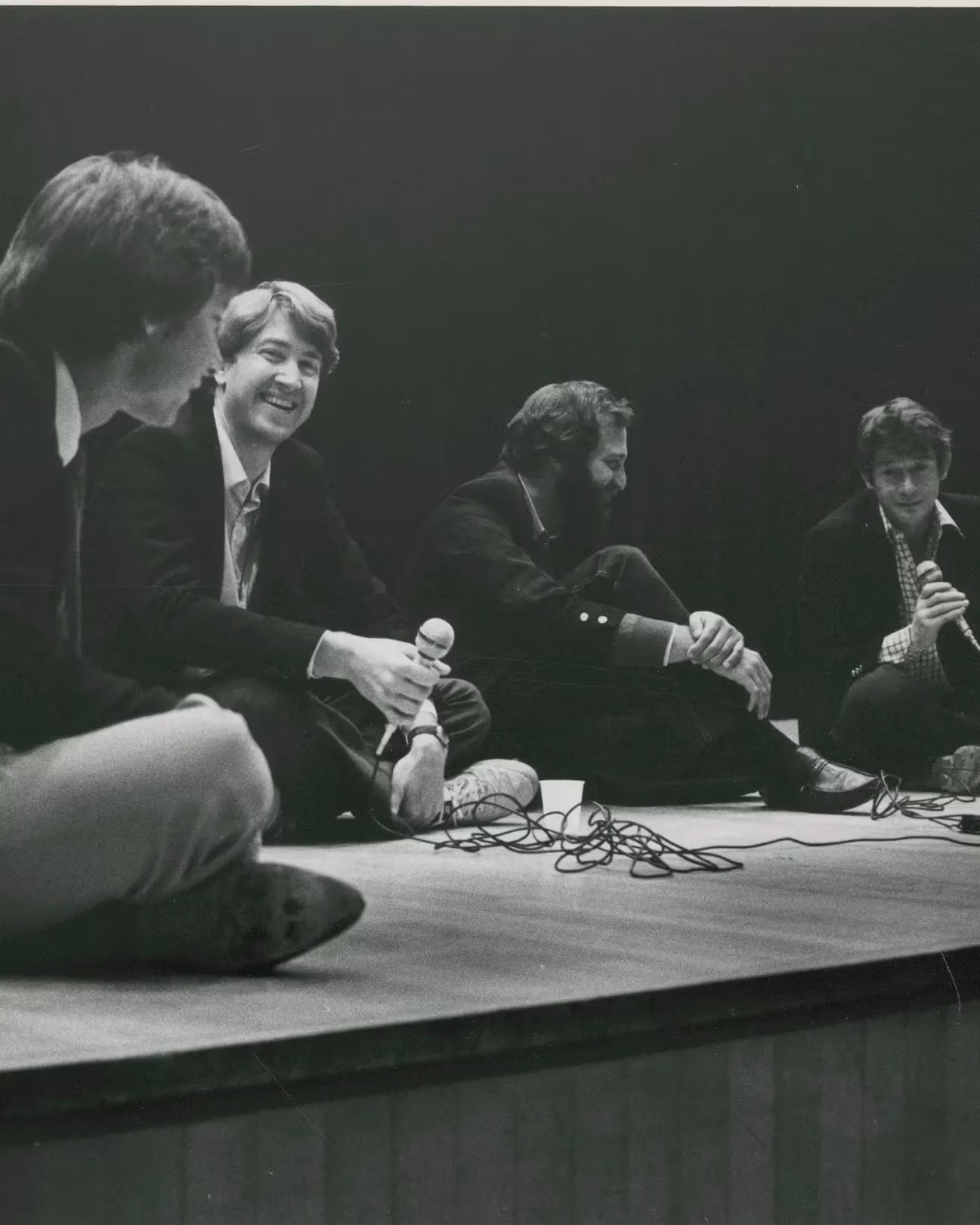 A black-and-white photograph of four men sitting on the floor of a stage, smiling and talking. Two of the men are holding corded microphones.