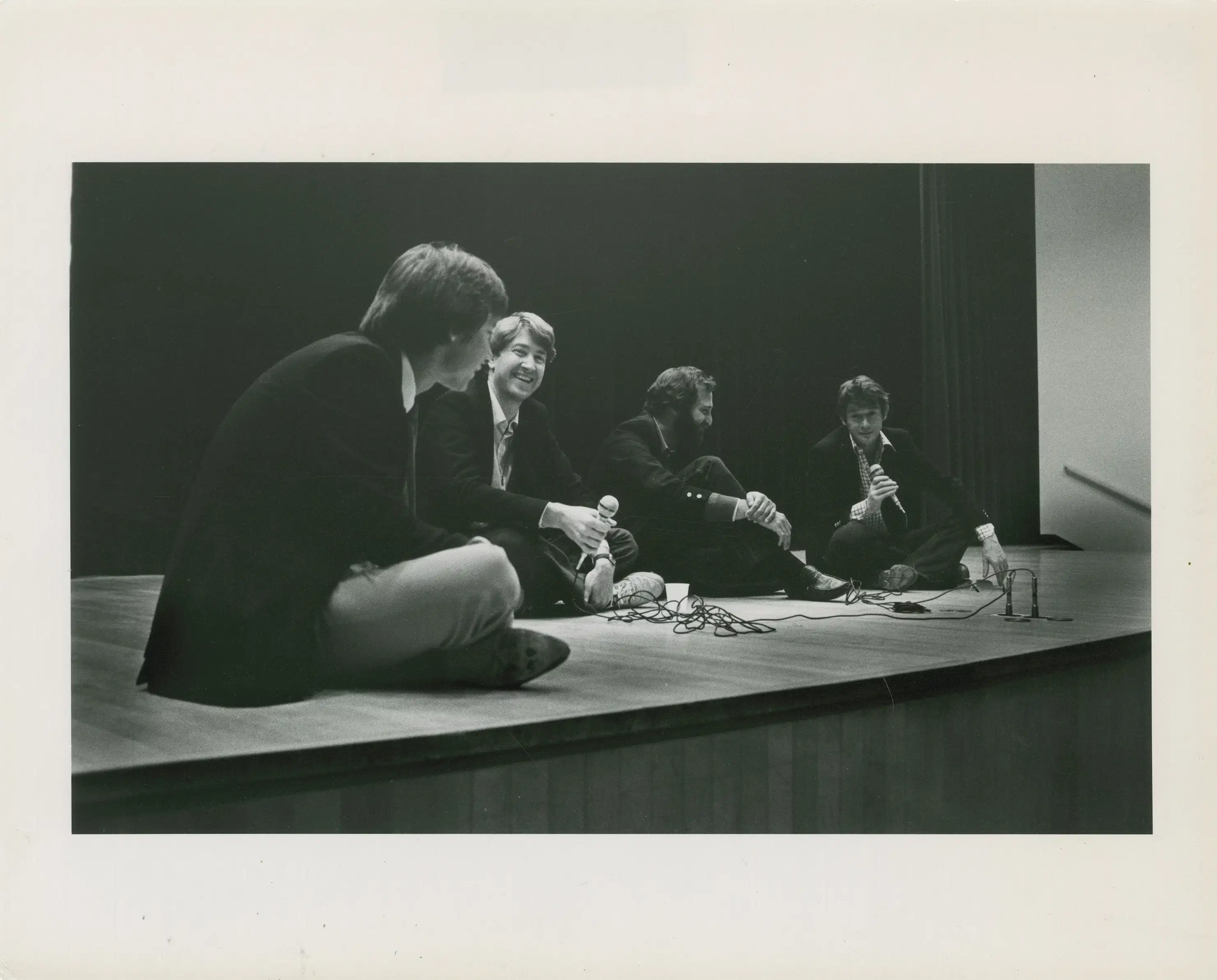 A black-and-white photograph of four men sitting on the floor of a stage, smiling and talking. Two of the men are holding corded microphones.