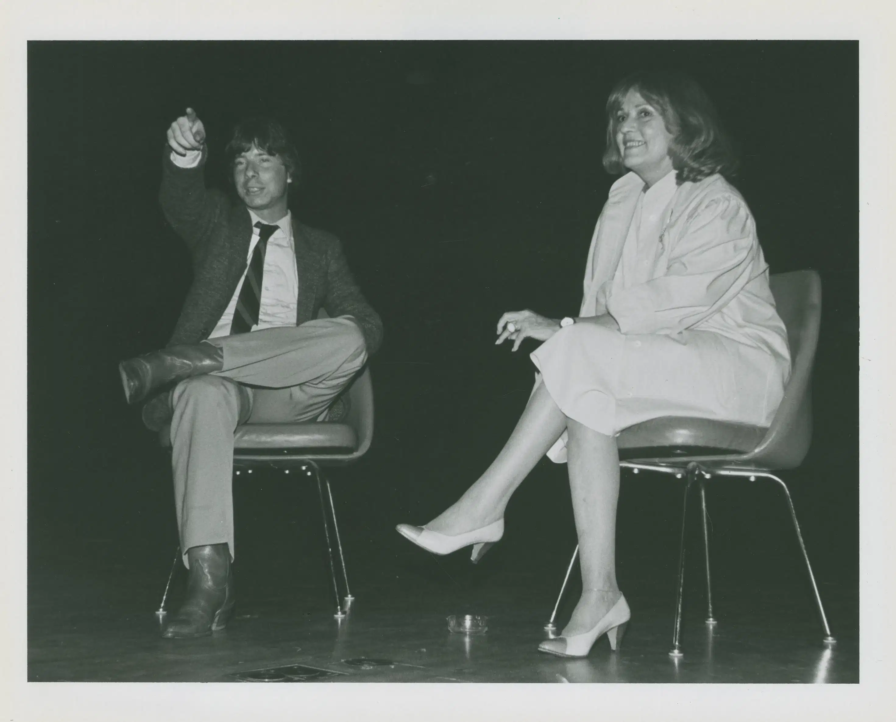 Two people sit in chairs on a stage in mid-discussion. The person on the left is pointing to something off-camera and both people are smiling.