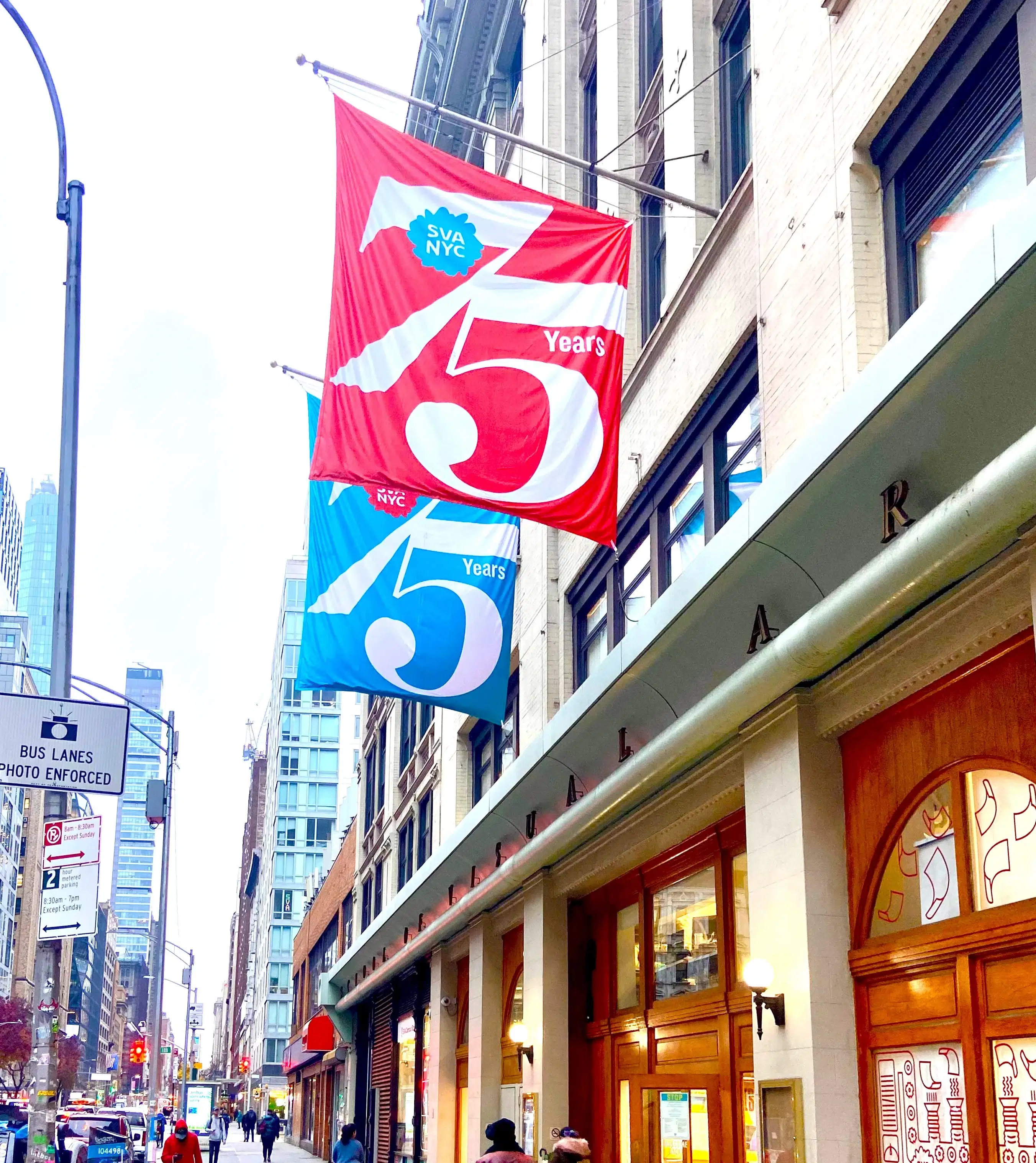Photograph of a New York City streetscape with the School of Visual Arts main building on the right side of the frame. Hanging outside the building are two flags, one red, one blue, that each say "75 Years" on them.