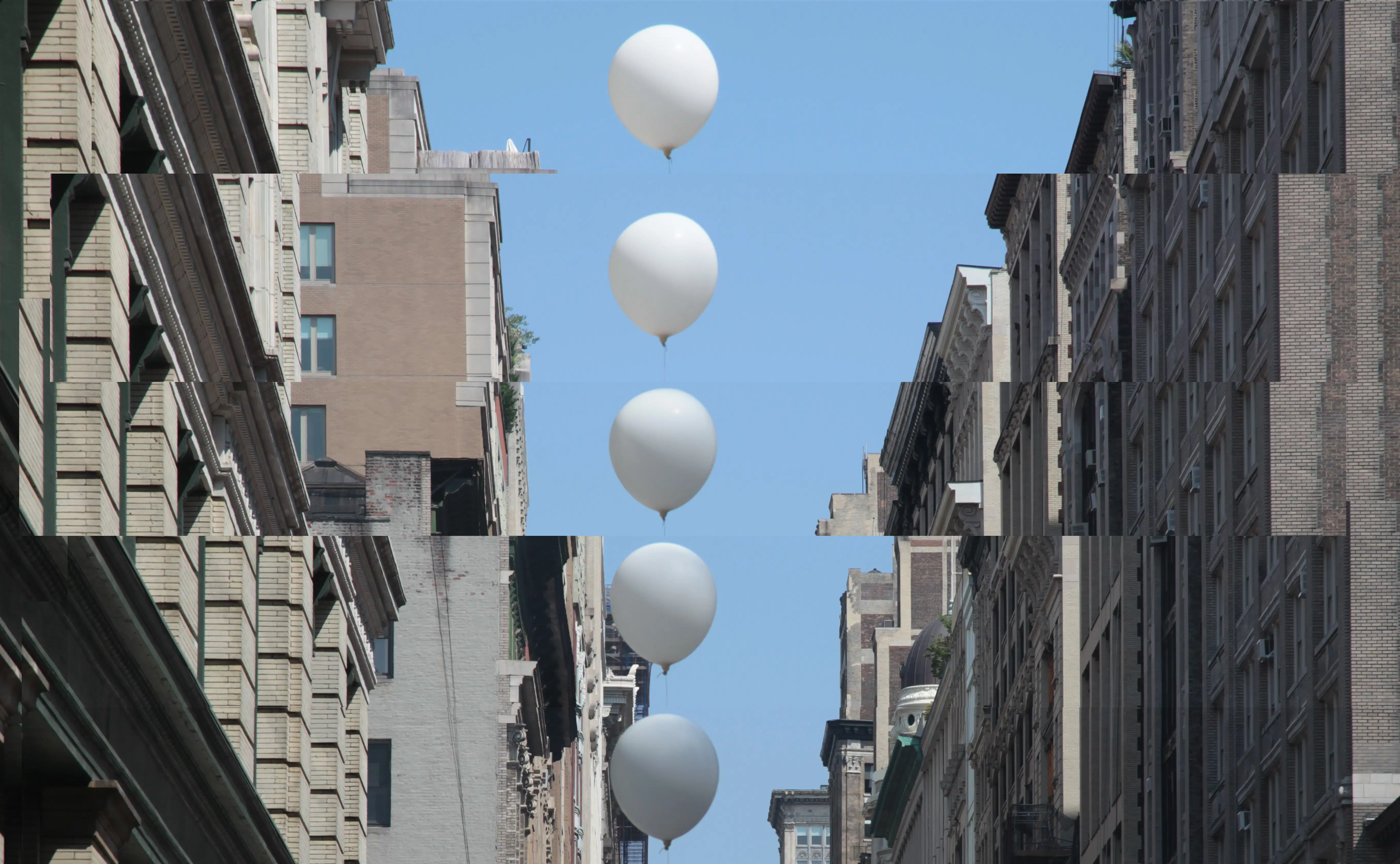A distorted photo of buildings on either side of the street, taken from below facing above so the viewer can see the buildings and a clear blue sky. A column of white balloons goes down the middle of the image.