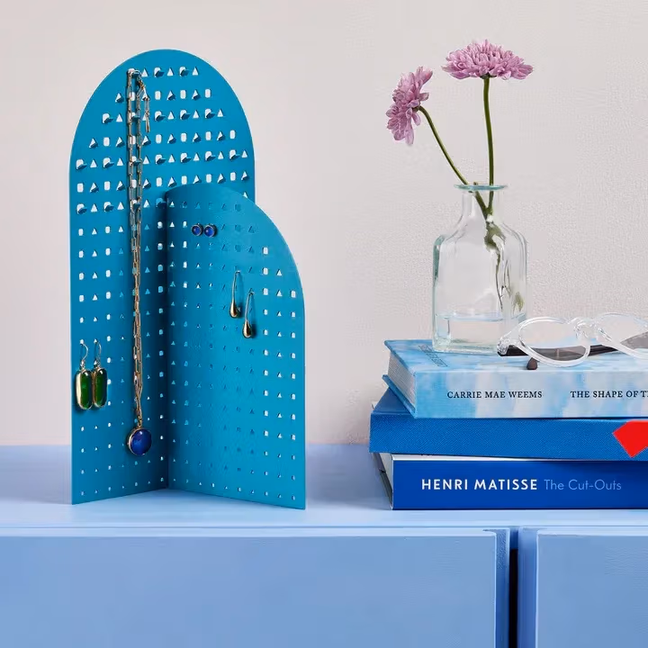 A blue jewelry stand made up of two arch shaped planes on a light blue table. There's also blue books and two flowers in a small glass vase.