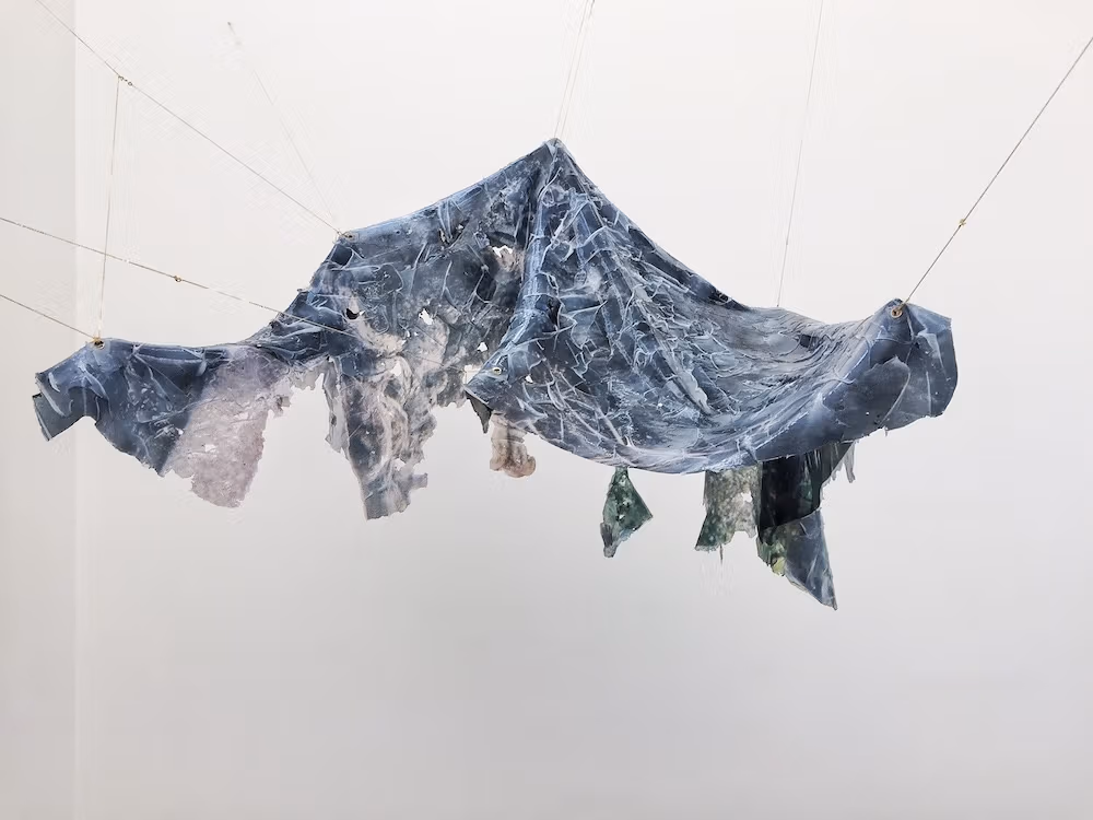 A dark piece of tarp suspended from the ceiling with wires floats in front of a white background