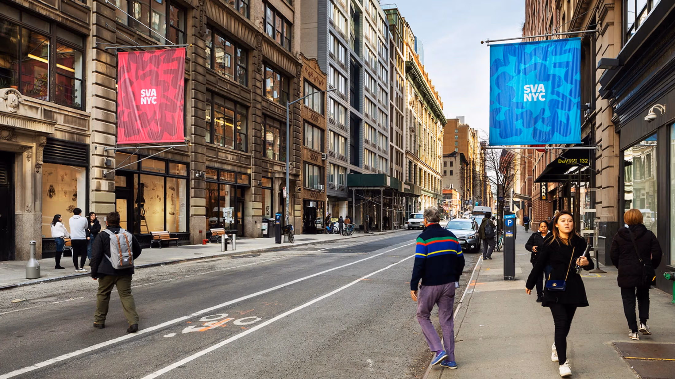 New York City street with pedestrians walking on both sides of the sidewalk as well as crossing the road; two large institutional flags are visible, the left being a red flag, flower outline and the words “SVA NYC,” the left being a blue flag, flower outline and the words “SVA NYC”