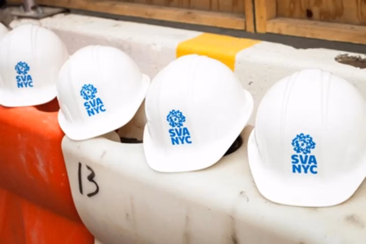 Four SVA NYC branded hard hats sit atop orange and white construction barriers