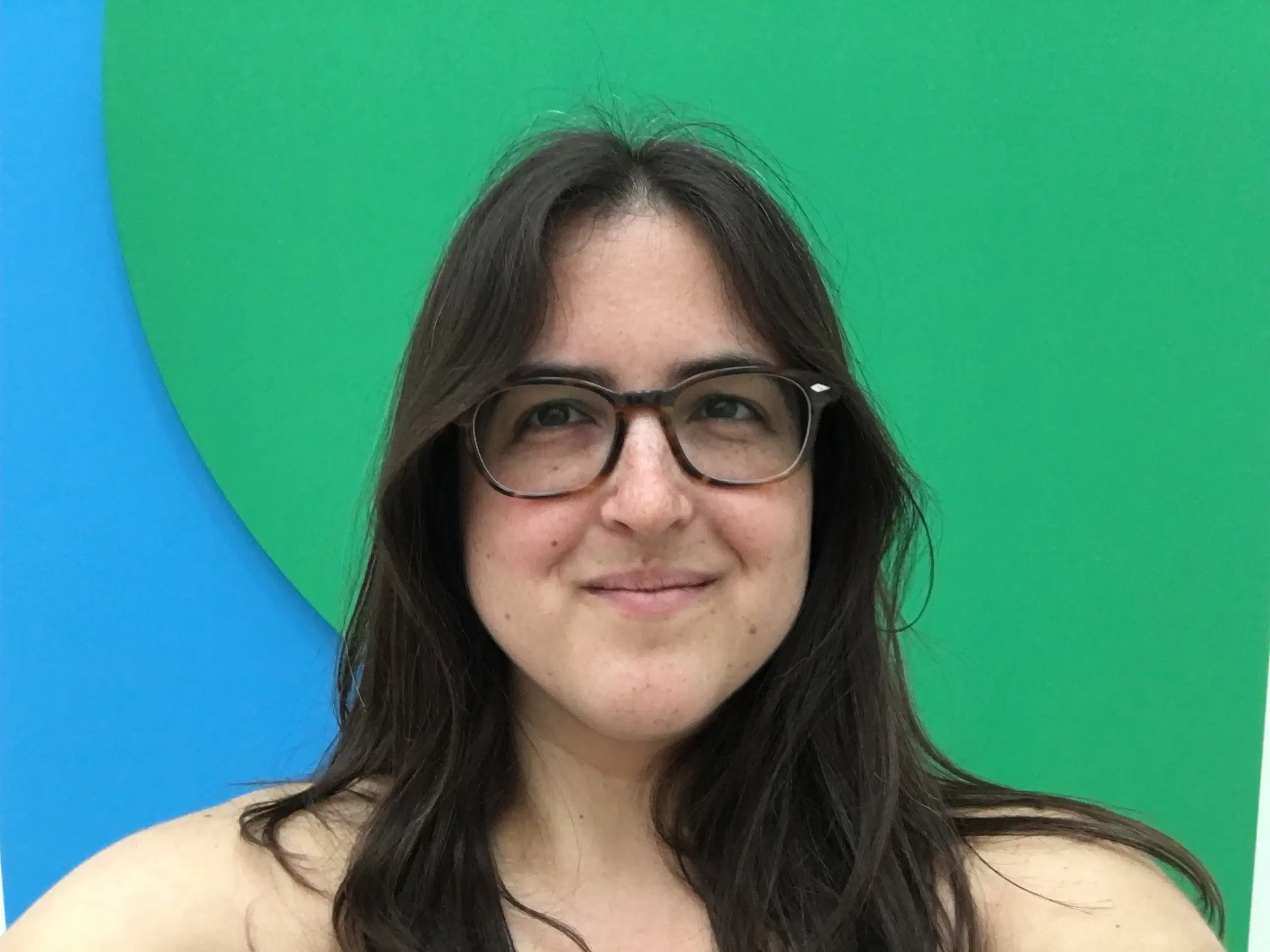 Curator Hayley Ferber smiles at the camera wearing glasses and standing in front of an abstract green and blue background.