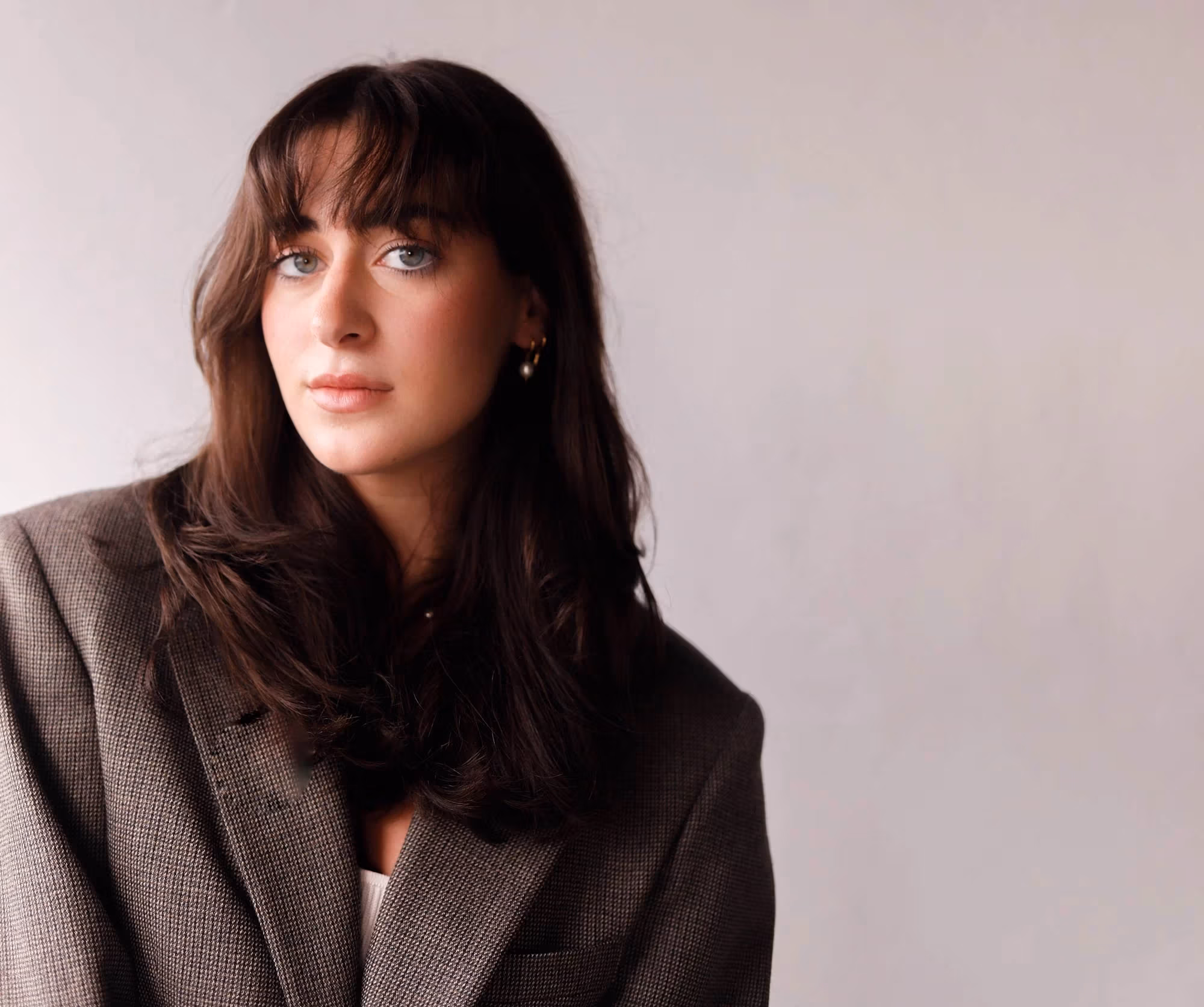 A student with brown hair and brown eyes wearing a grey suit jacket looking at the camera with a grey background.