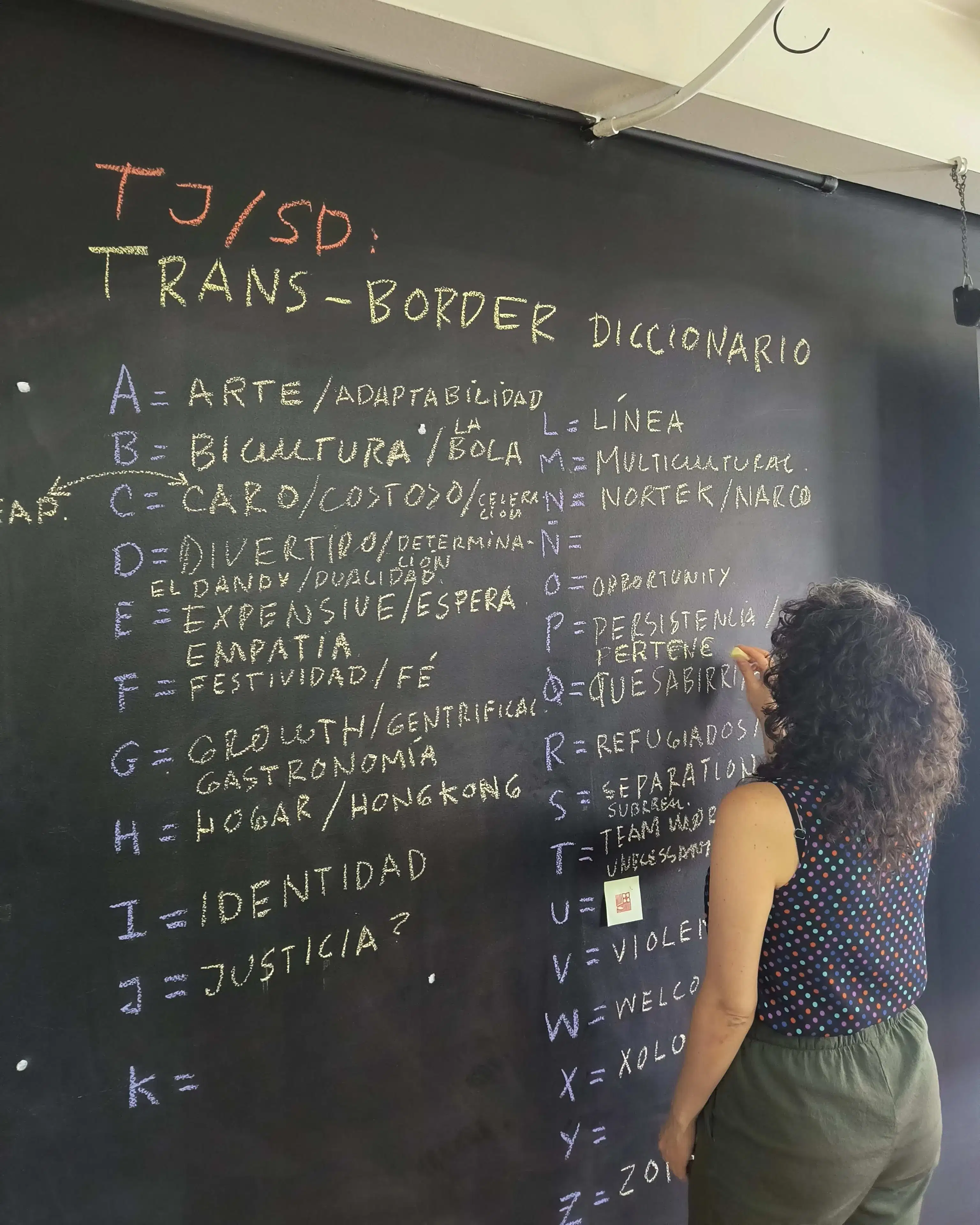 A woman writes Spanish words in yellow chalk on a blackboard next to letters of the alphabet written in purple chalk.