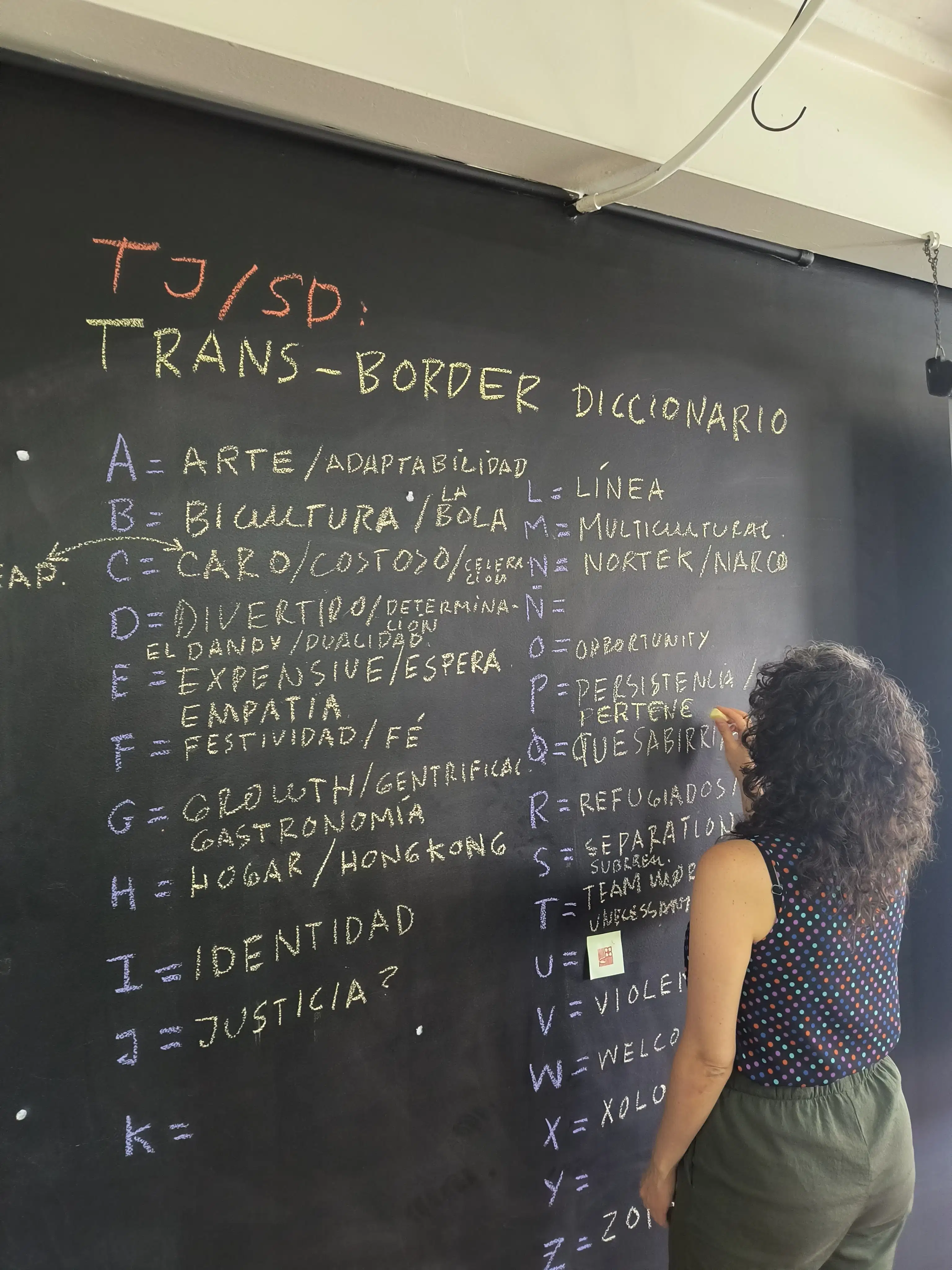 A woman writes Spanish words in yellow chalk on a blackboard next to letters of the alphabet written in purple chalk.