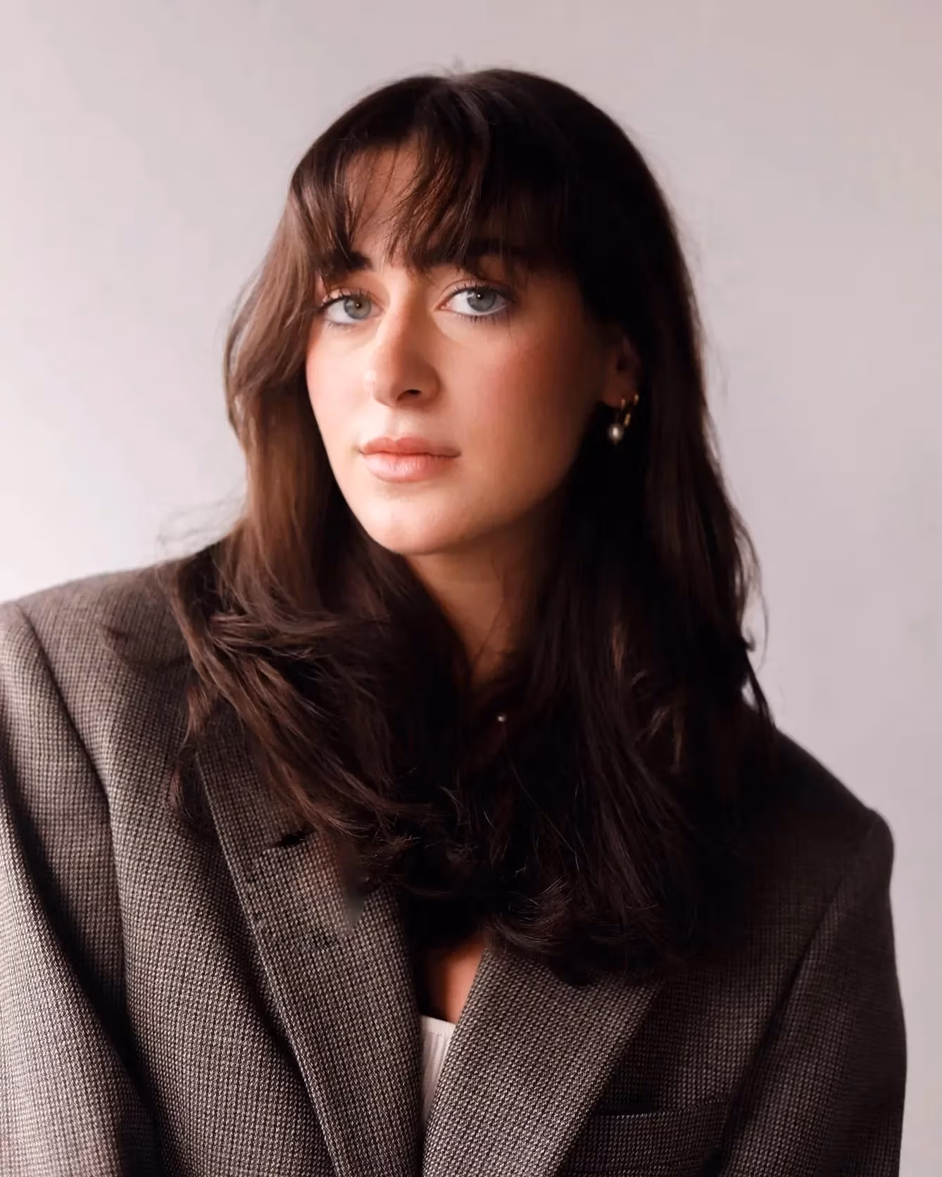 A student with brown hair and brown eyes wearing a grey suit jacket looking at the camera with a grey background.