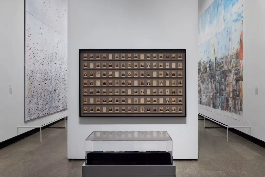 a gallery with large paintings hung on the walls and a standalone wall in the center displaying rows of small photographic works 