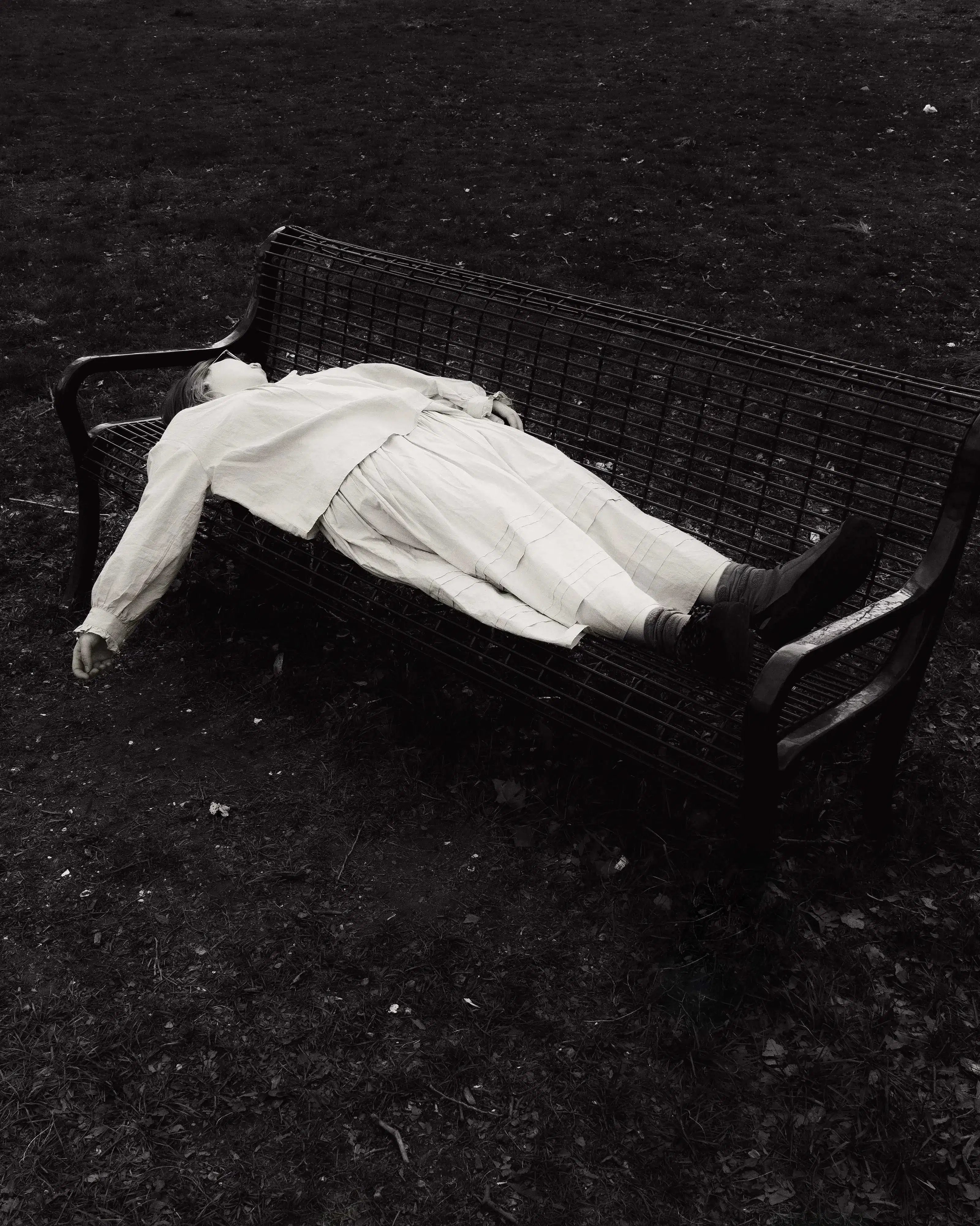 A black and white photograph of a girl lying on a bench, her hand hanging loosely, face obscured. The bench and her body are surrounded by chaotic bundles of grass.