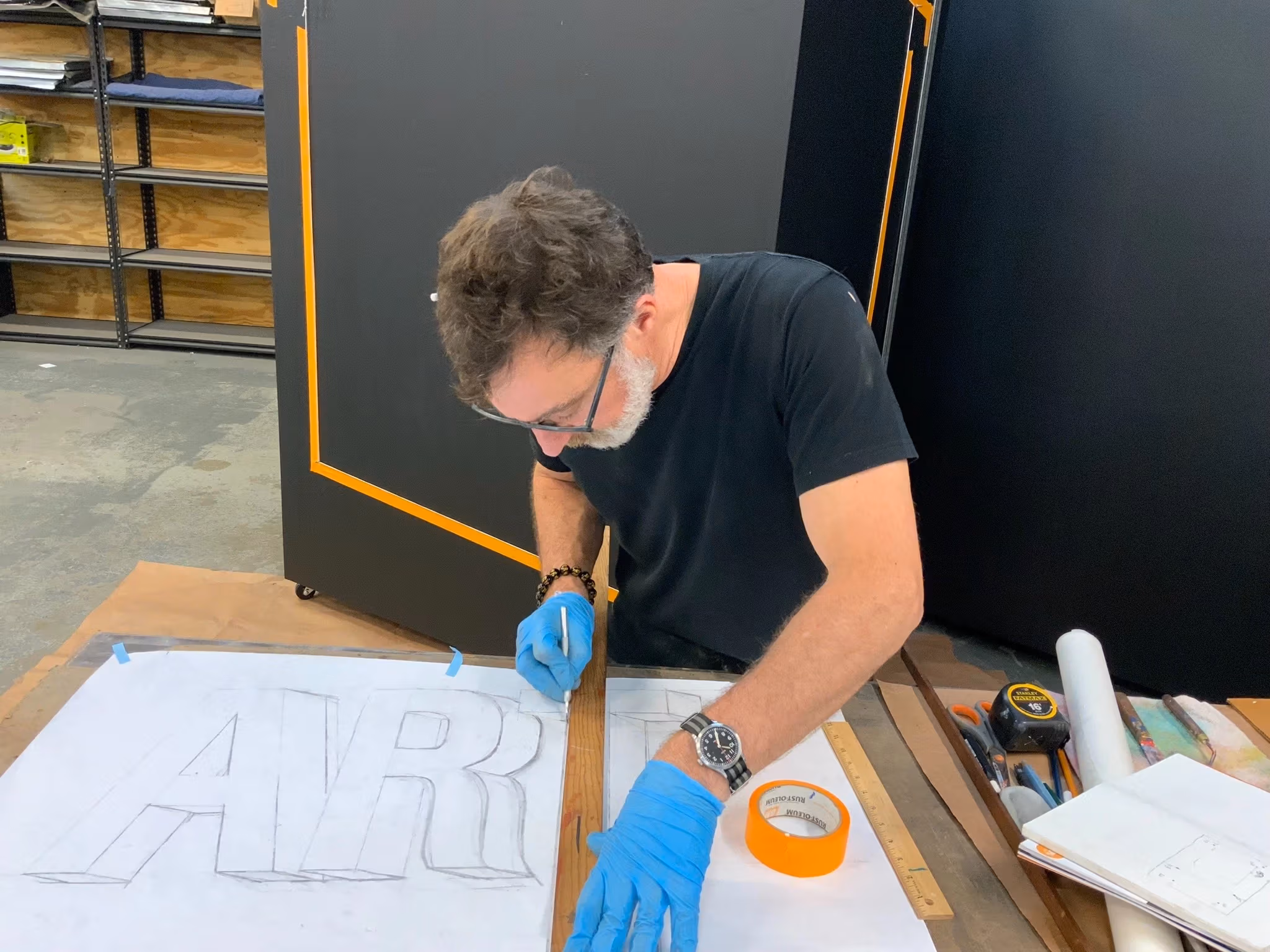 A man, stephen gaffney, working in his studio on a drawing that reads "AR.." on top of a wooden worktable.
