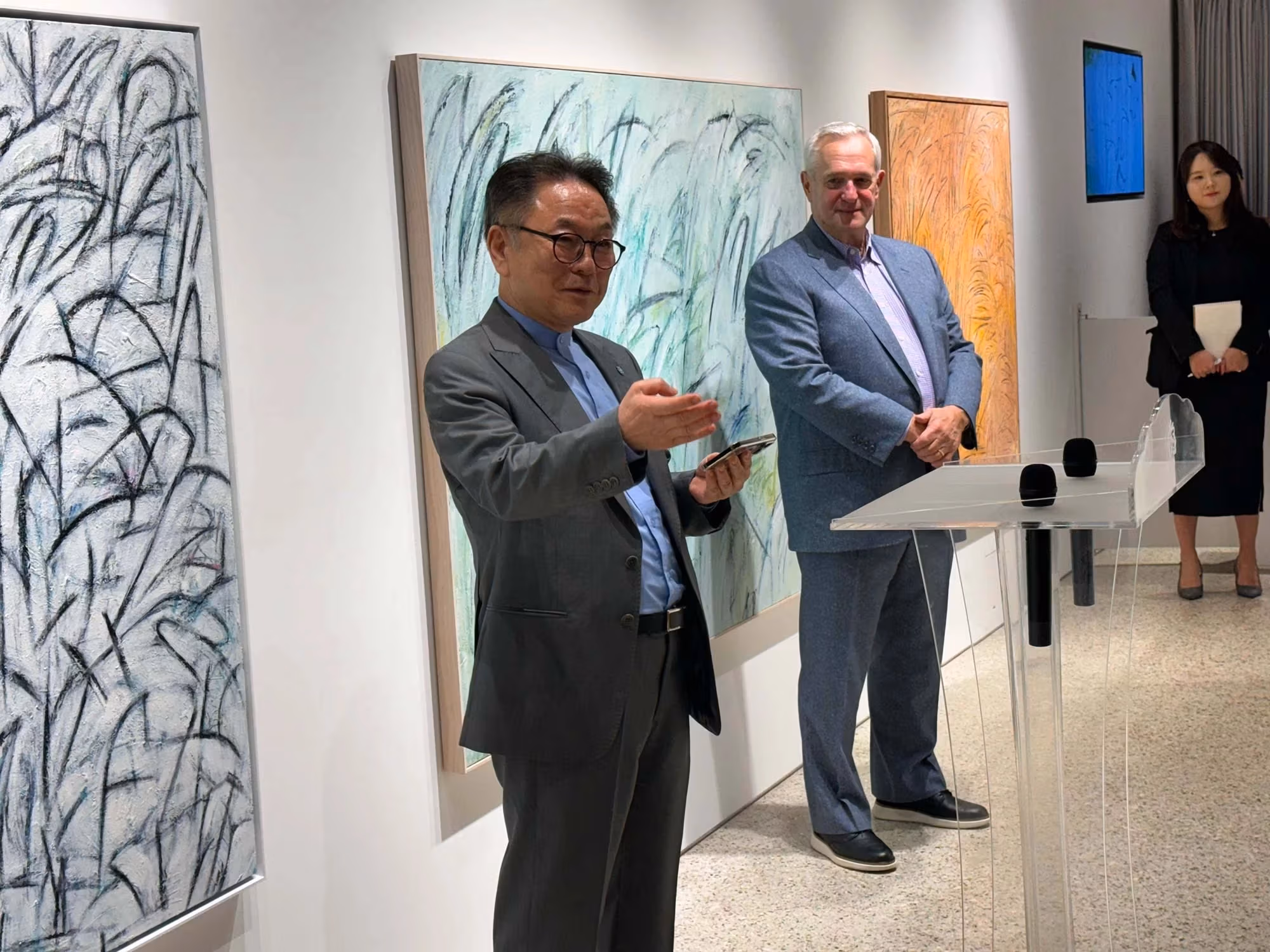 A man in a gray suit holding a phone gestures towards an audiences as another man with white hair in a similar, but lighter suit stands nearby smiling. 