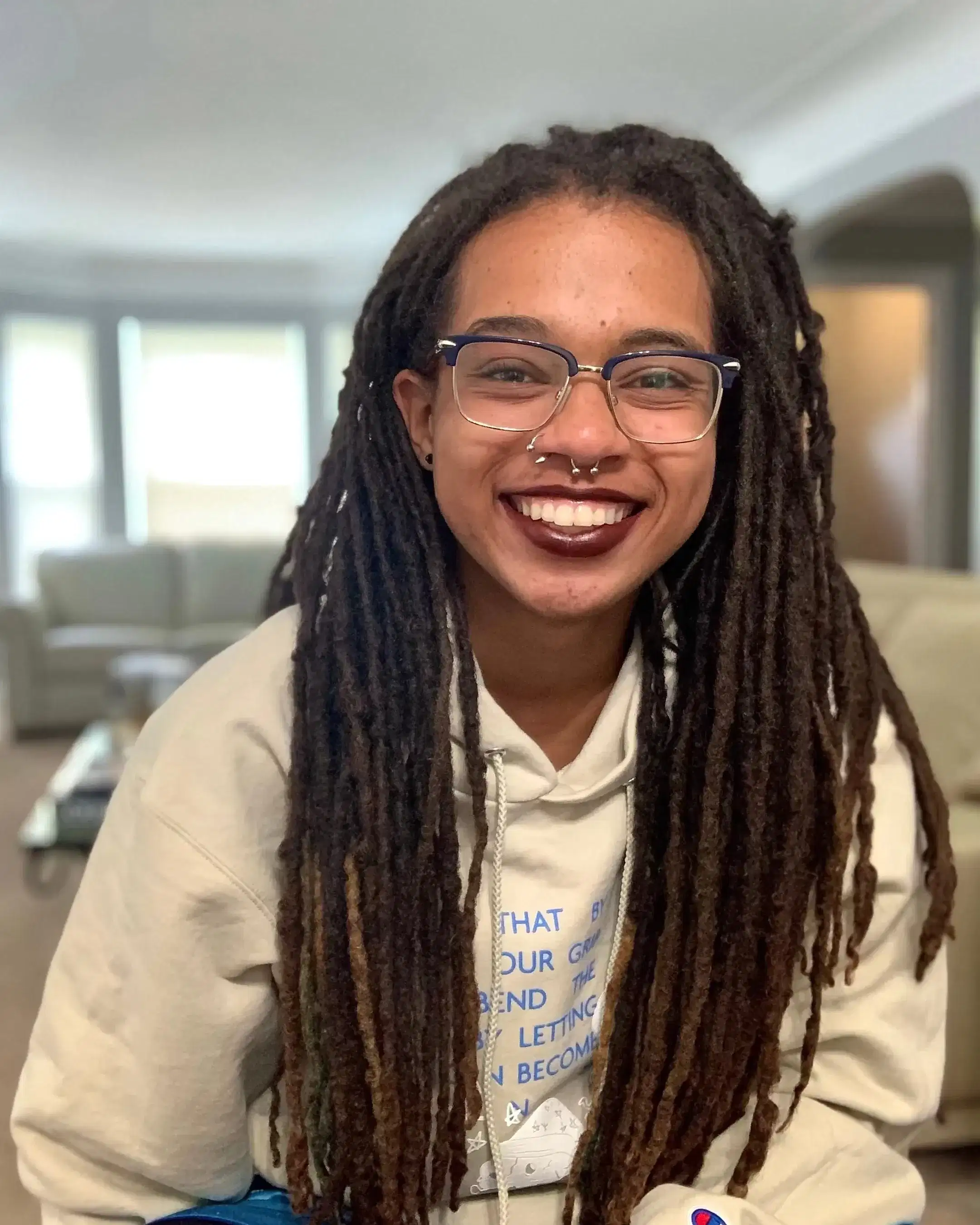 An individual with glasses, nose piercings, dark lipstick, and locs smiles at the camera warmly.