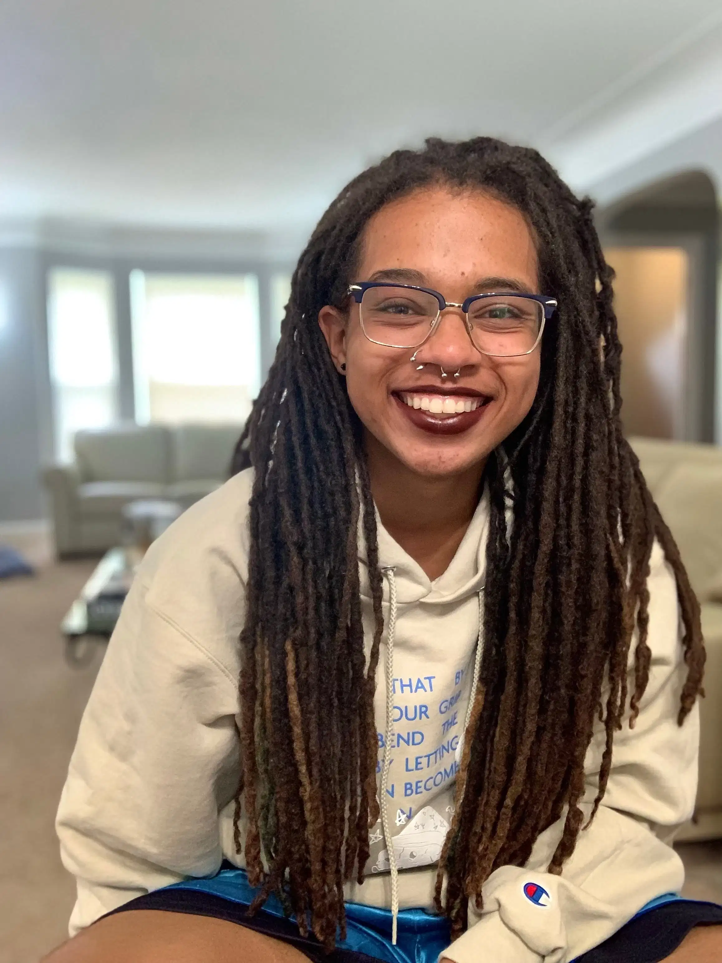 An individual with glasses, nose piercings, dark lipstick, and locs smiles at the camera warmly.