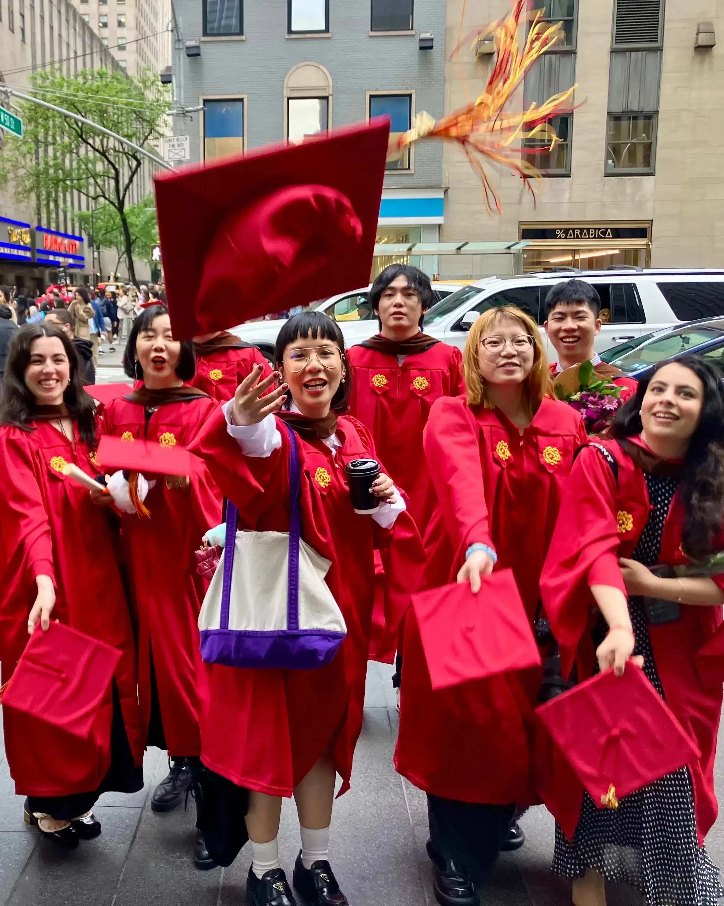 The Masters in Digital Photography graduating class of 2025, in the iconic SVA red graduation gowns, in front of the famed Radio City Music Hall where the commencement ceremony just took place. The students are throwing their caps in excitement. 