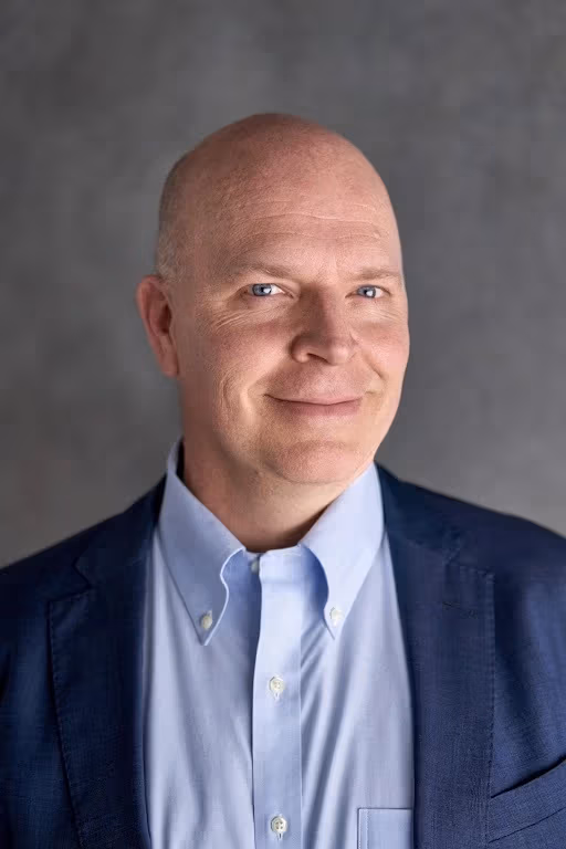 A man in a sky blue collared shirt and darker blue suit jacket smiles into 