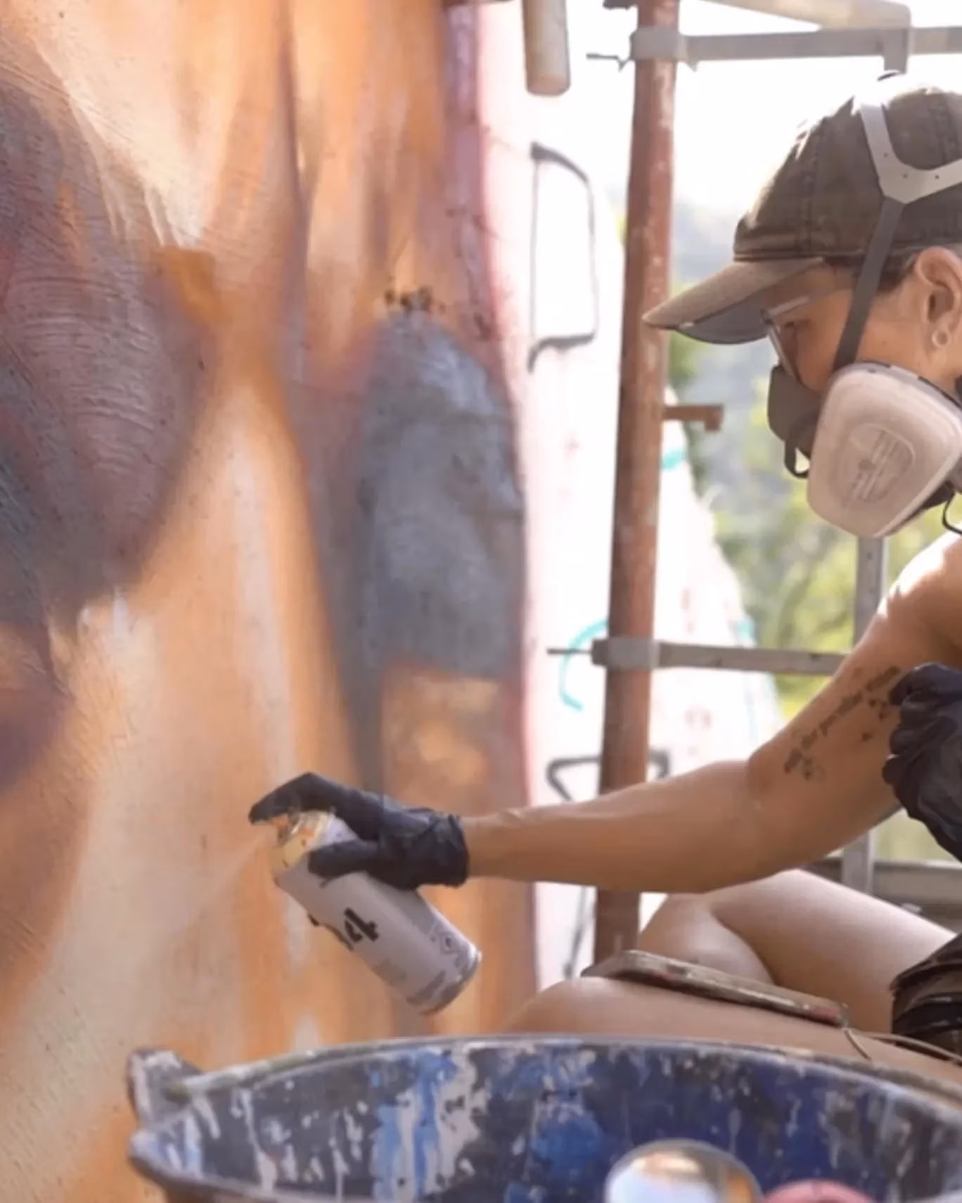 A woman wearing a hat and respirator spray painting a large mural of someone's face.