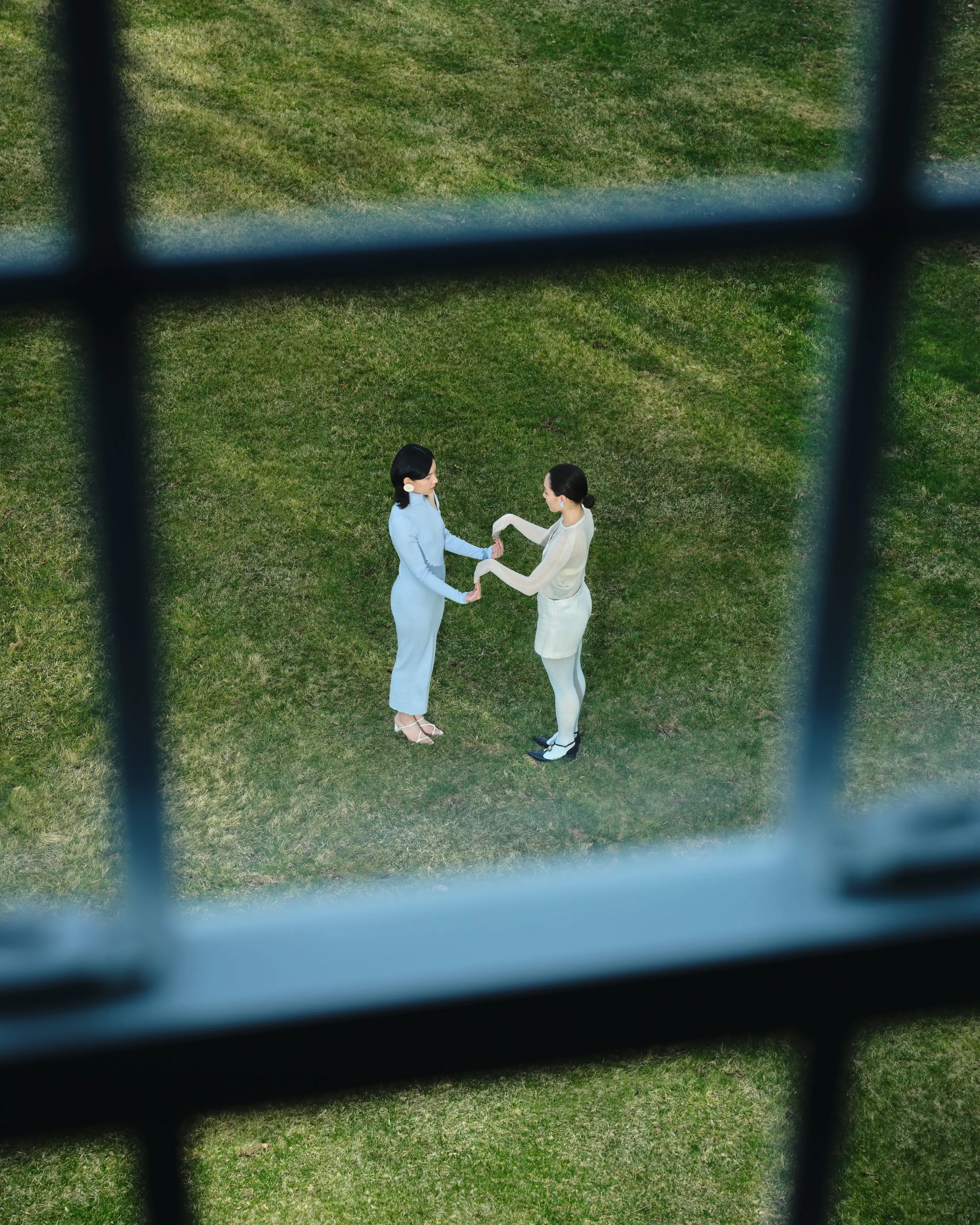  Two women stand face-to-face on a green lawn, gently holding each other’s hands, seen through a window grid from above.