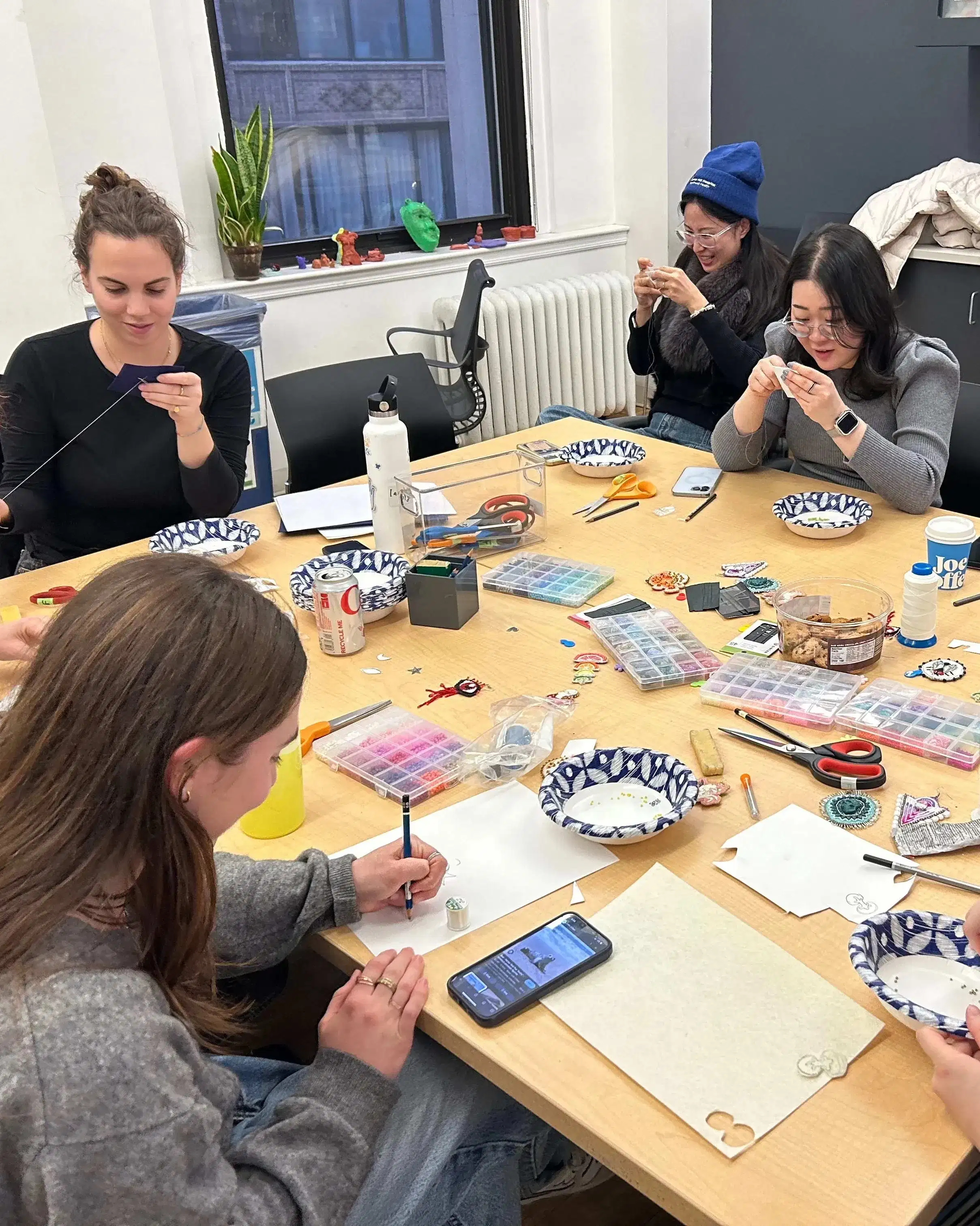 students gathered around a square wood table covered in art supplies working on a beading project 