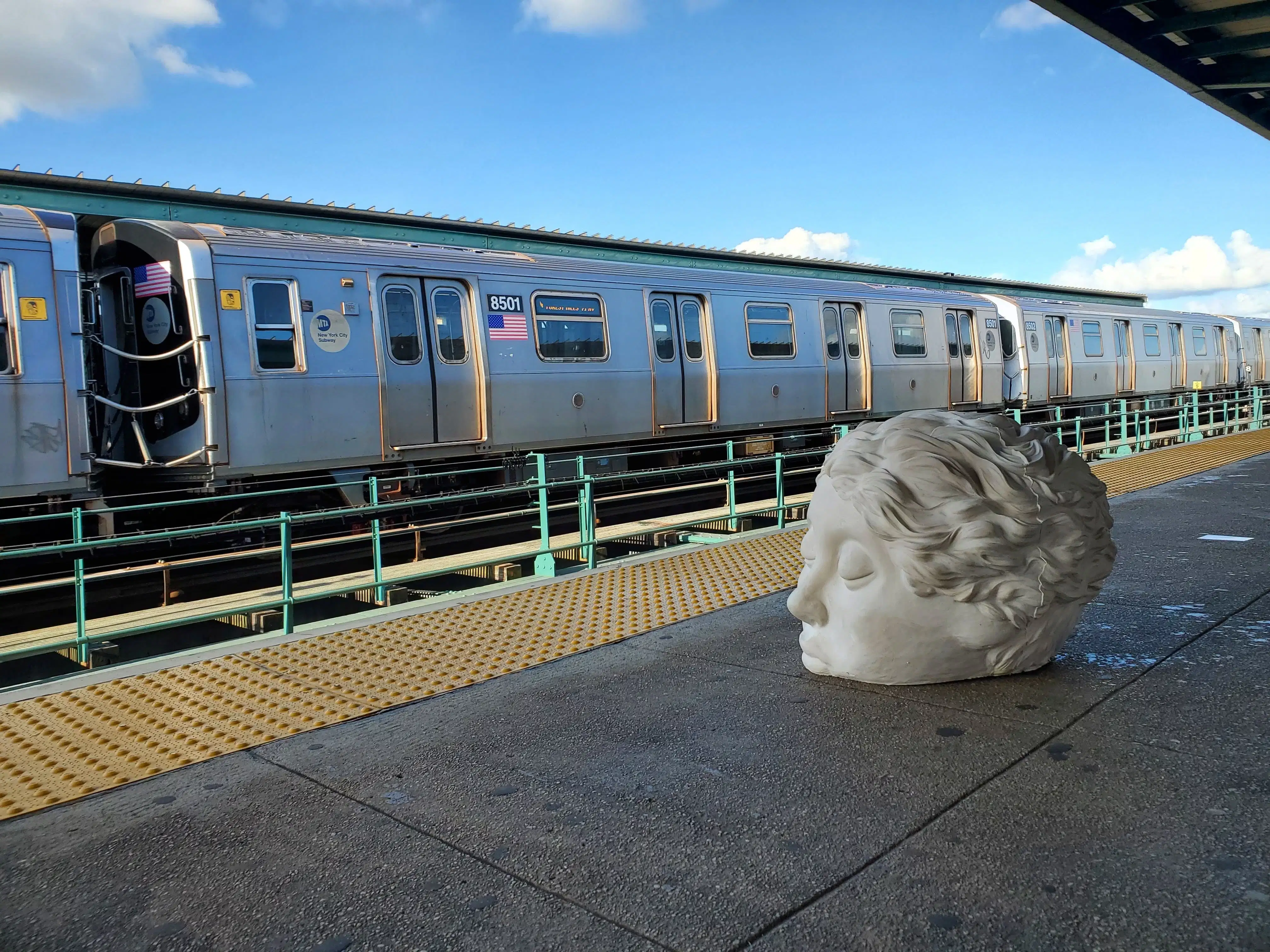 Large sculpture of a head on an above ground subway platform facing a train