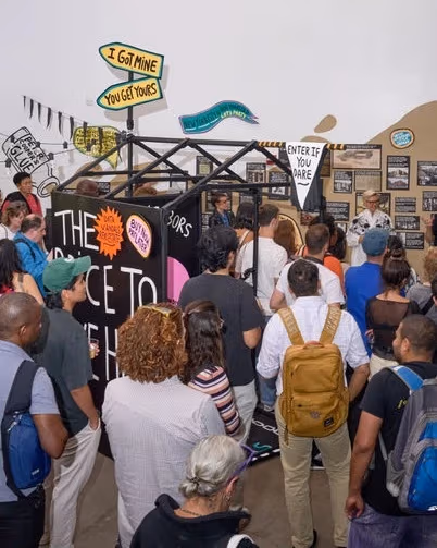A crowd of people stands in a gallery space, gathered around a central art installation with colorful protest-style signs on the walls.