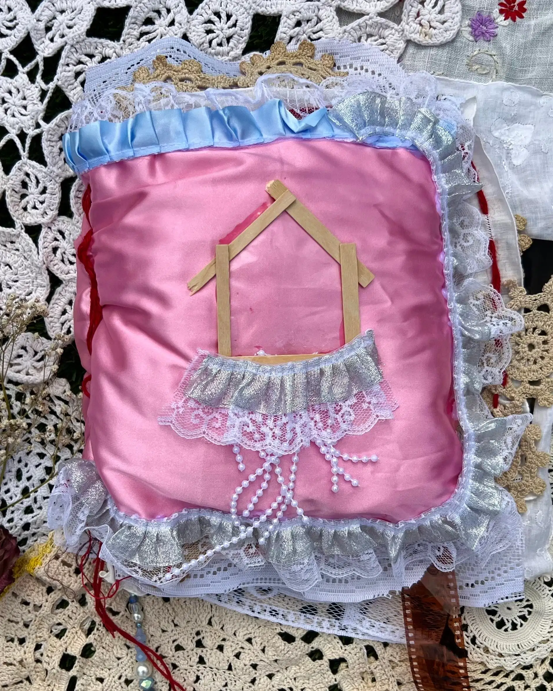 soft pink satin square pillow book cover with image of house made of light wooden sticks with thick strip of white lace and thin strips of threaded beads hanging down. Blue lace is sewn into the top of the cover. A film strip is seen at the bottom of the pillow. This pillow book is placed on pieces of white and off-white antique like lace.