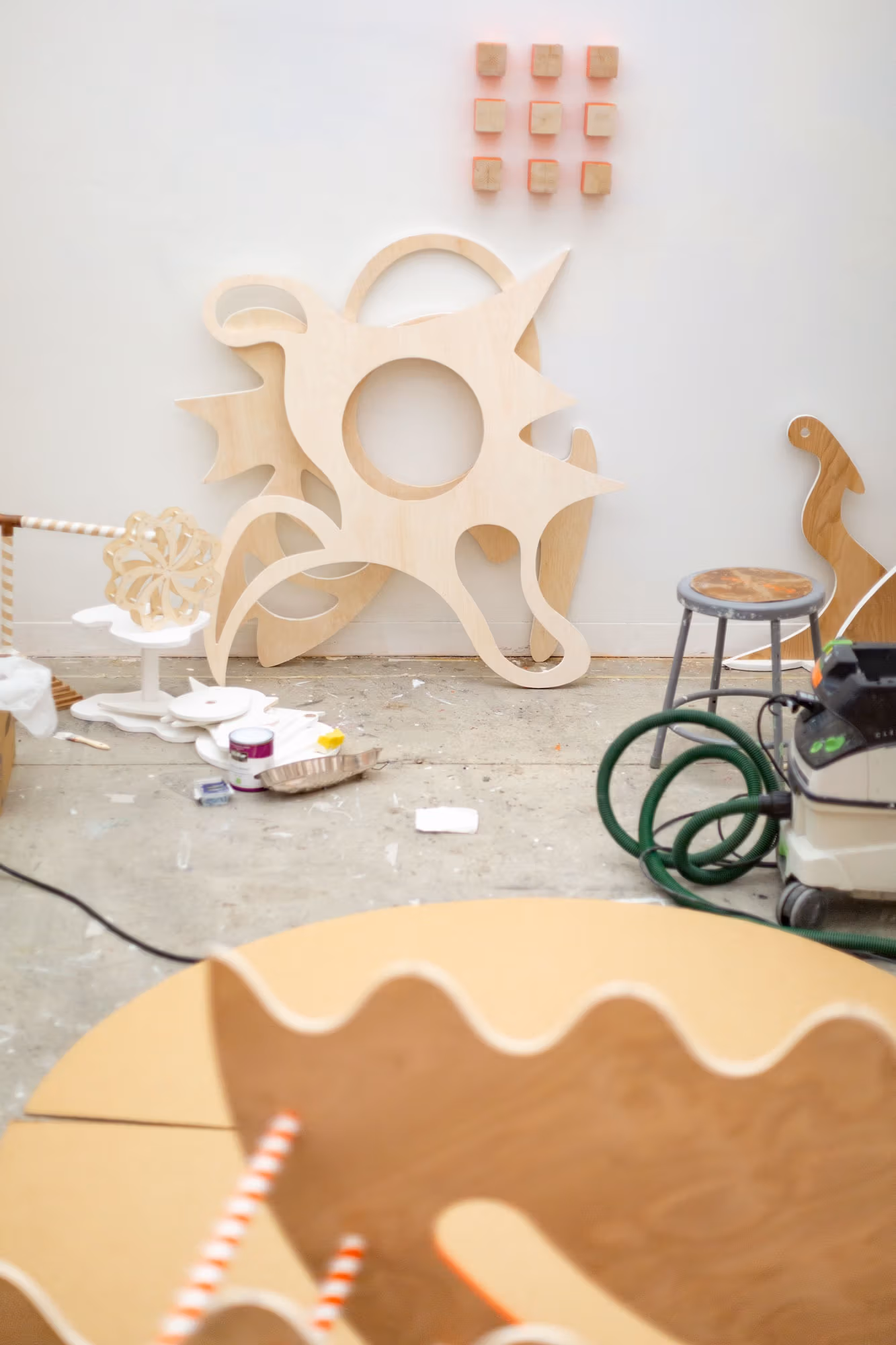 An installation view of Miley Huang's studio. There are wooden sculptures resting against the wall with some small square wood pieces hanging on the right side. On the ground there are miscellaneous items, such as paint, paint tray, chair, and vacuum. In the foreground is another wooden sculpture.