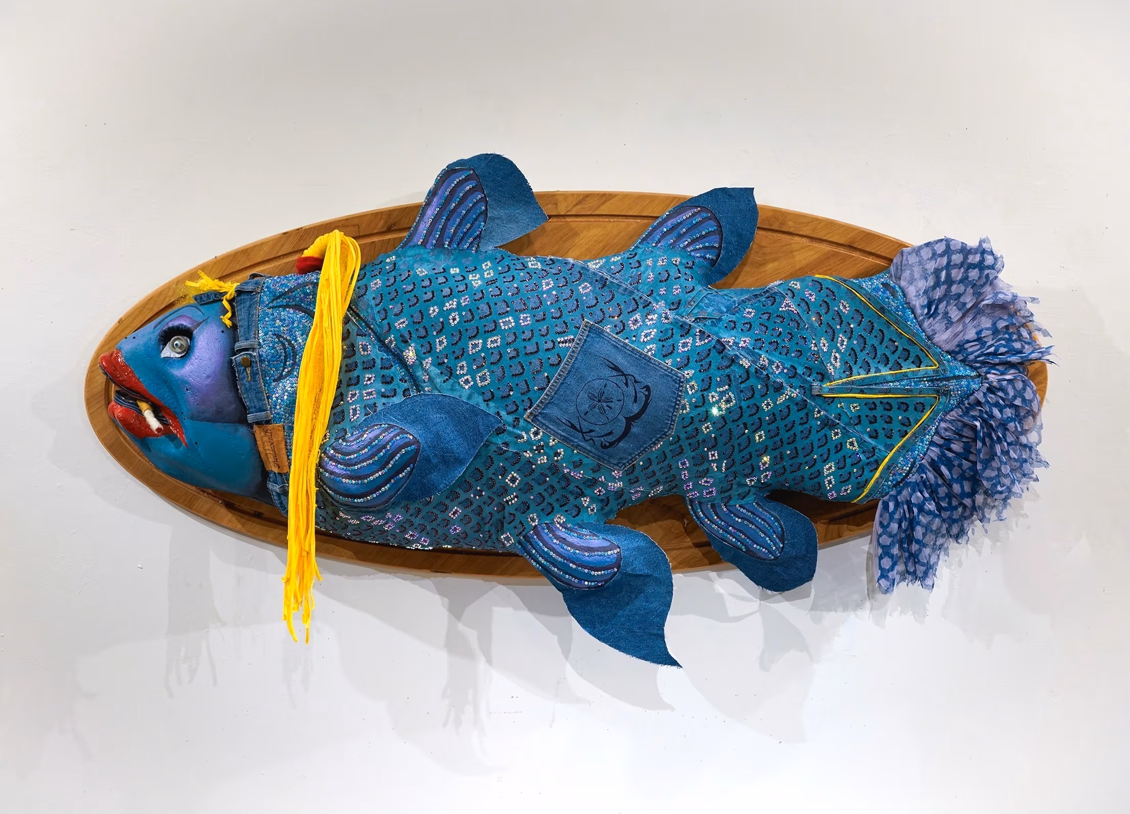 Artistic fish sculpture on a wooden plaque features vibrant blue fabric, yellow yarn, and a patch, blending whimsy with a unique textile design.