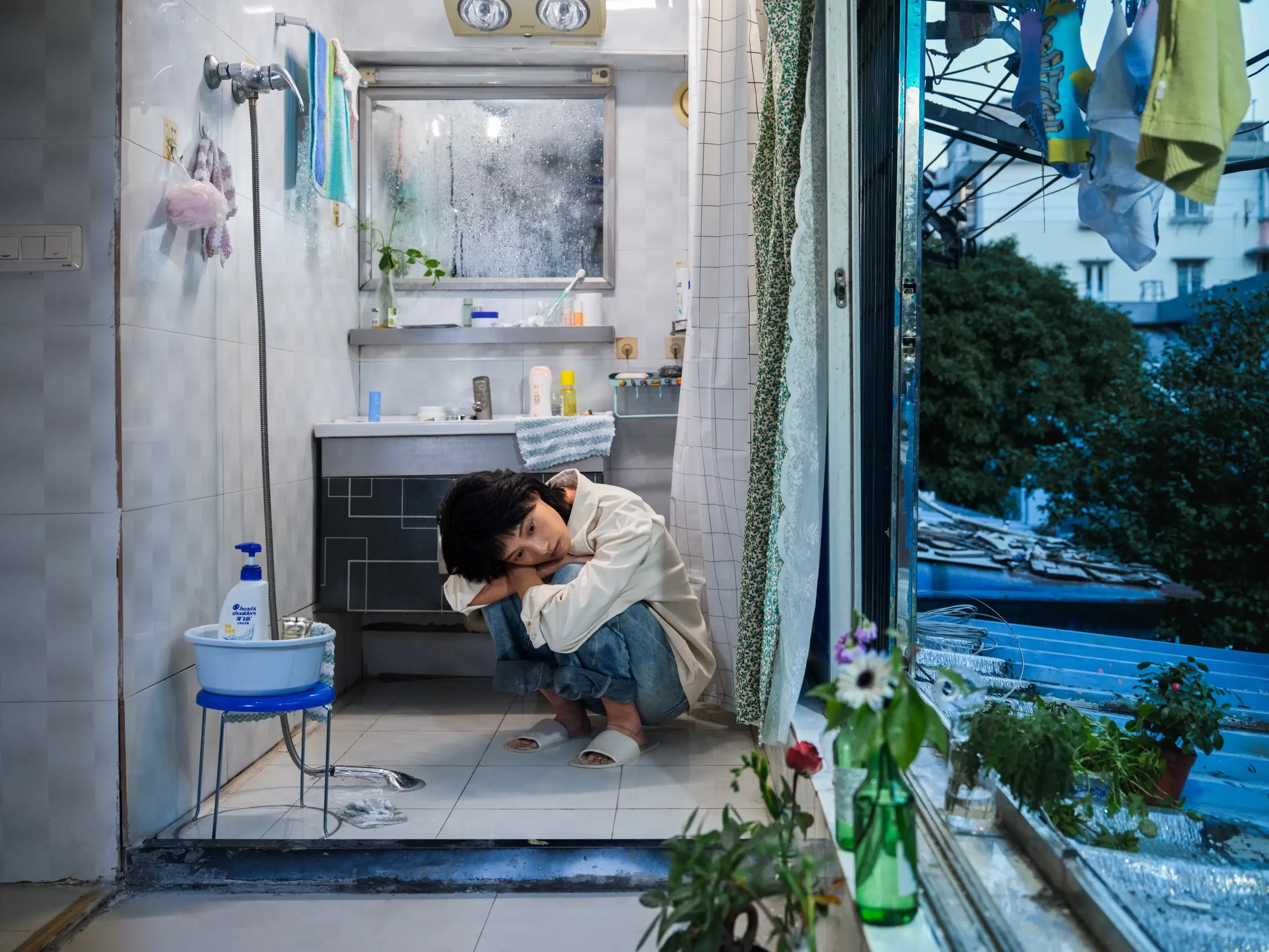 A color photography by Shaohan Fang entitled En Route. In a dim bathroom at dusk, the girl crouches on a wet floor, staring at the window. Water pools around her feet, she appears visibly overwhelmed.
