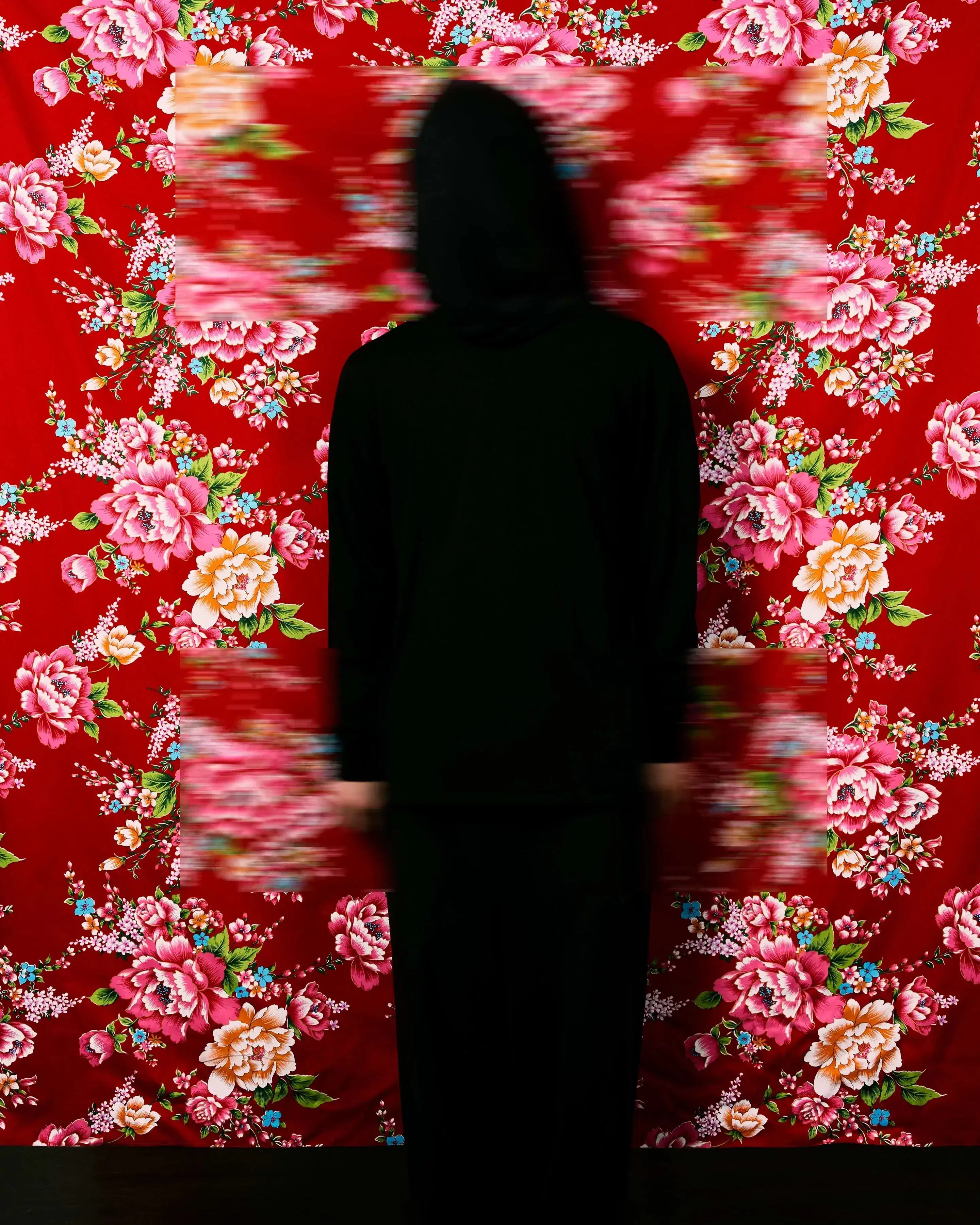 A color photograph featuring a figure wrapped in black from head to toe standing facing the camera against a red Hakka floral fabric background, which is suspended and reveals a faint dark wooden floor below. Three blurred rectangles cover the head, hands, and feet.