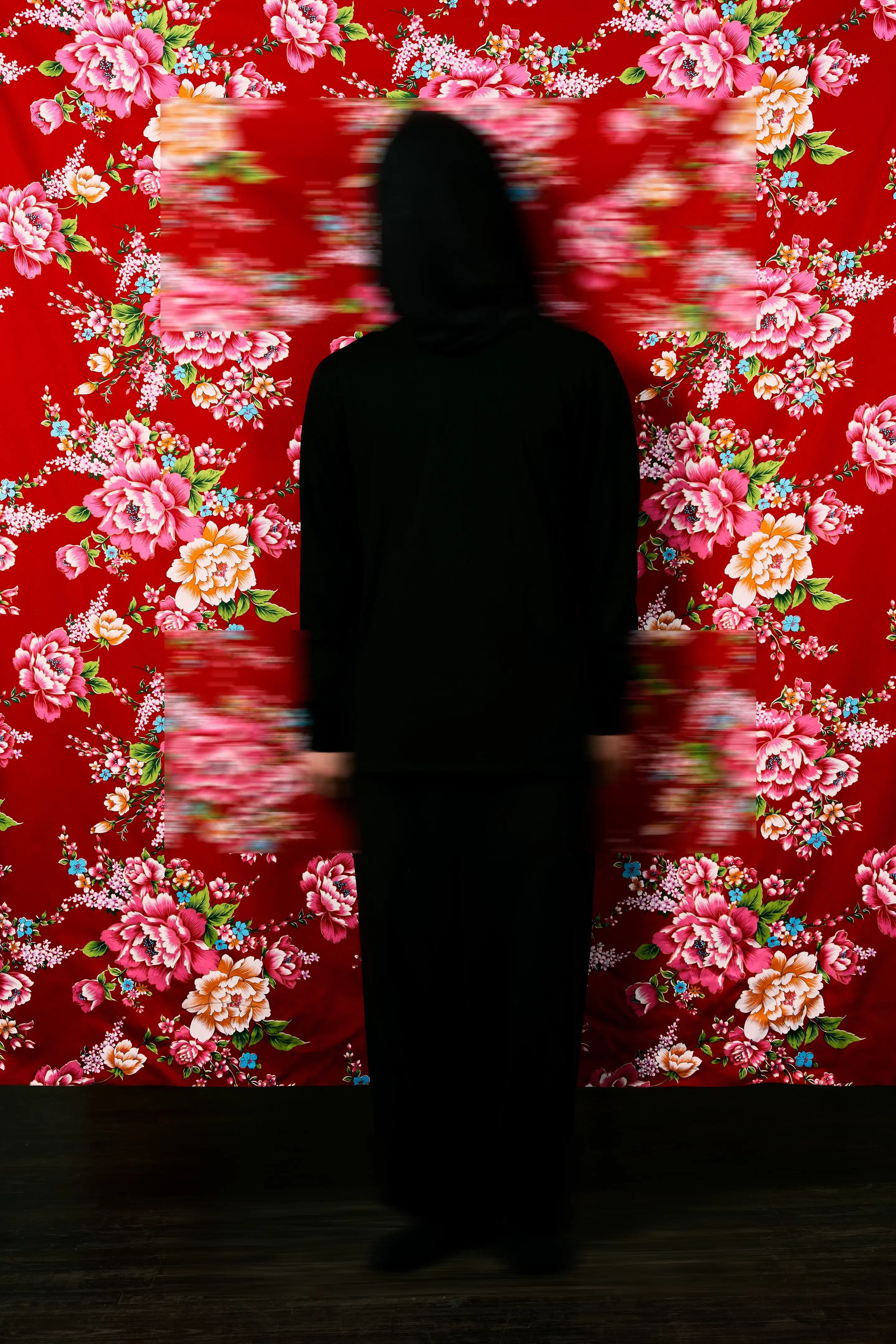 A color photograph featuring a figure wrapped in black from head to toe standing facing the camera against a red Hakka floral fabric background, which is suspended and reveals a faint dark wooden floor below. Three blurred rectangles cover the head, hands, and feet.