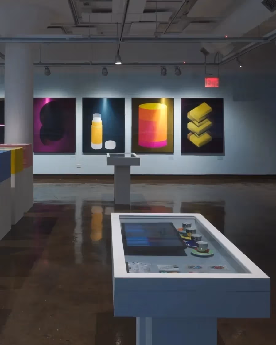 Wide view of an art exhibition in situ at SVA Chelsea Gallery including work hung on walls, vitrines and large stacks of ephemera for visitors to take.